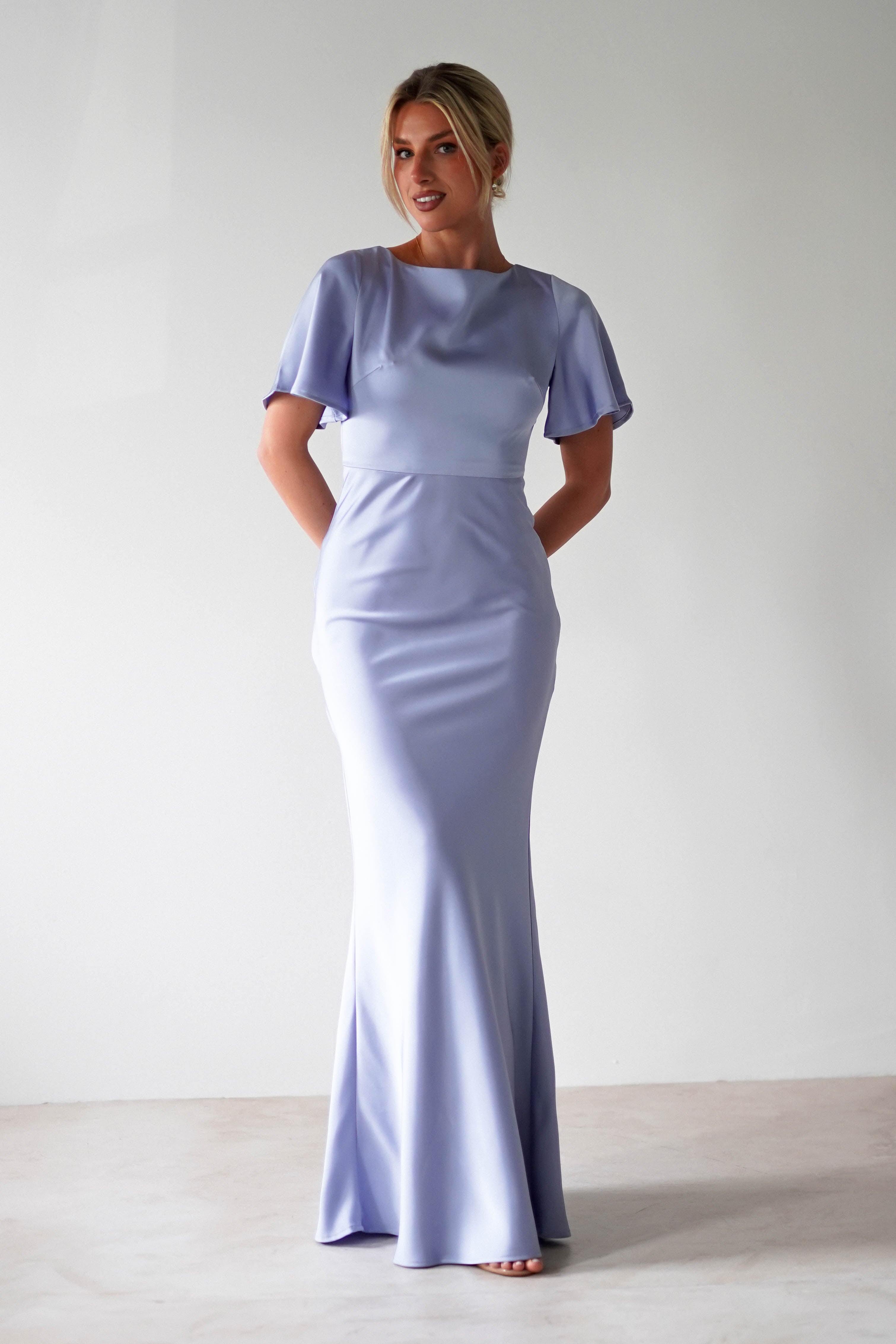 A woman in a light purple satin maxi dress stands barefoot against a plain light background, smiling with her hands behind her back.