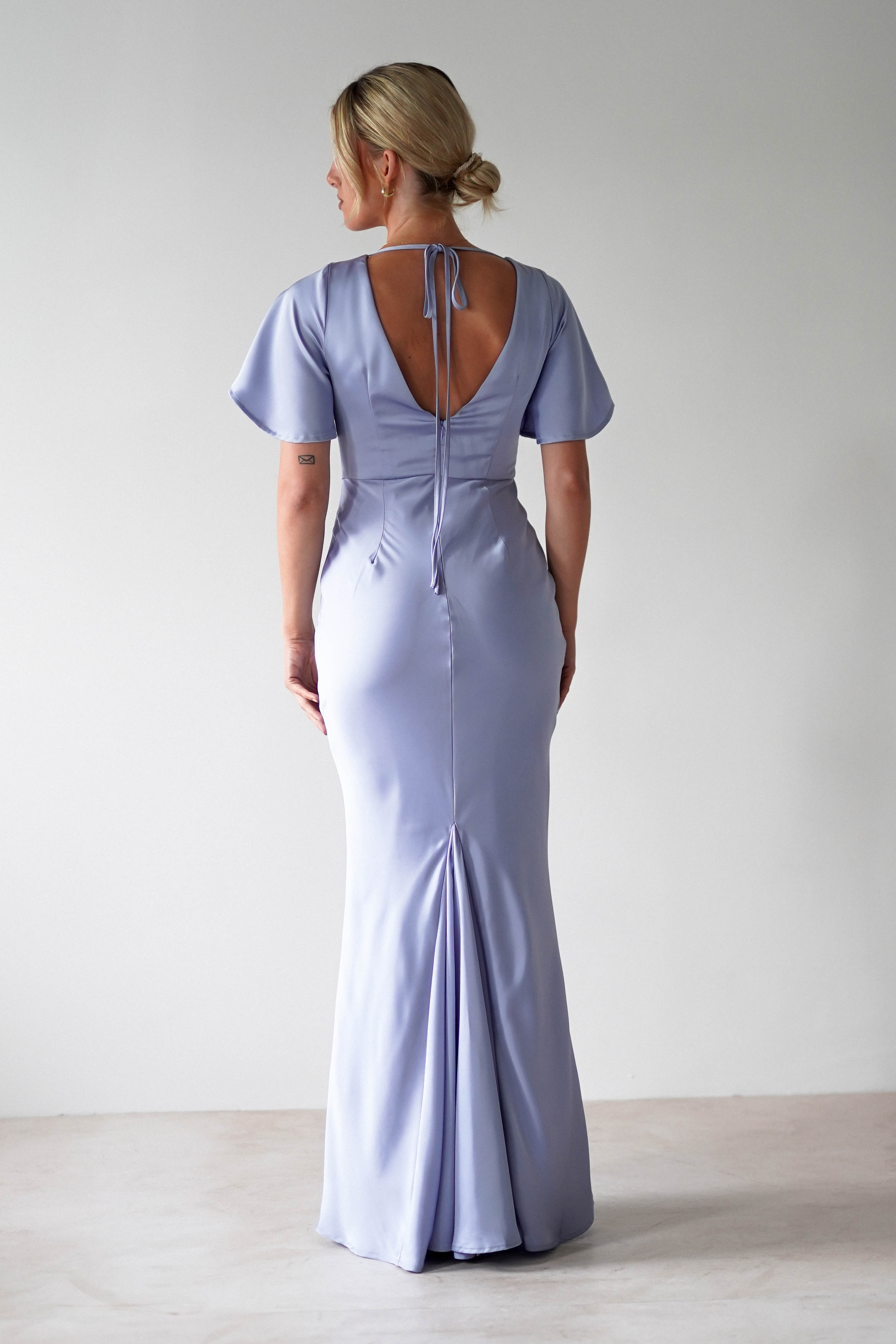 A woman stands facing away, wearing a light lavender satin maxi dress with short flutter sleeves, a fitted waist, an open back with a tie, and seam detailing—an elegant option for a wedding guest dress. Her blonde hair is styled in an updo.