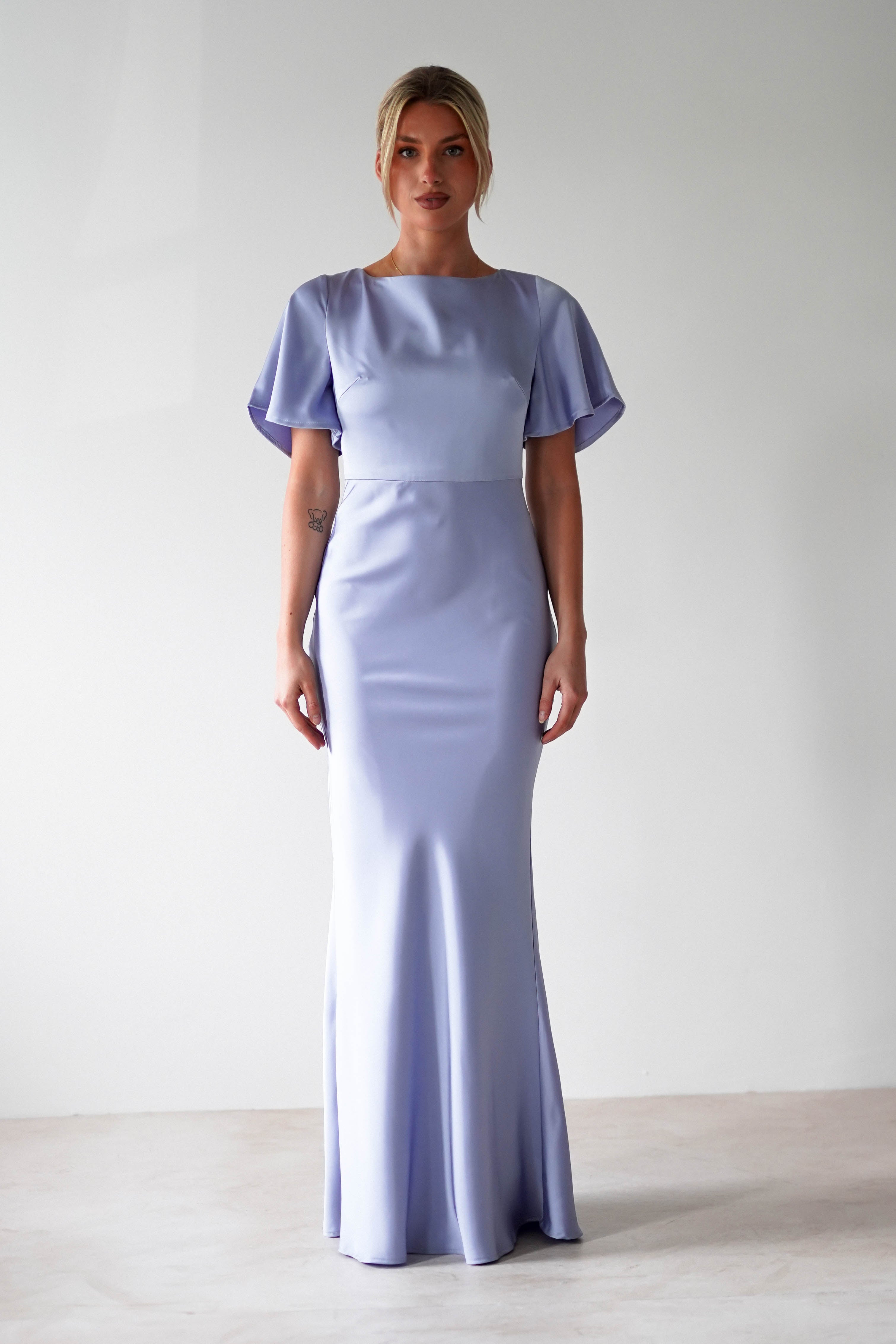 A woman stands against a plain background wearing a floor-length, light lavender satin maxi dress with short flutter sleeves and a fitted silhouette—perfect as a bridesmaid dress or for any elegant wedding guest look.