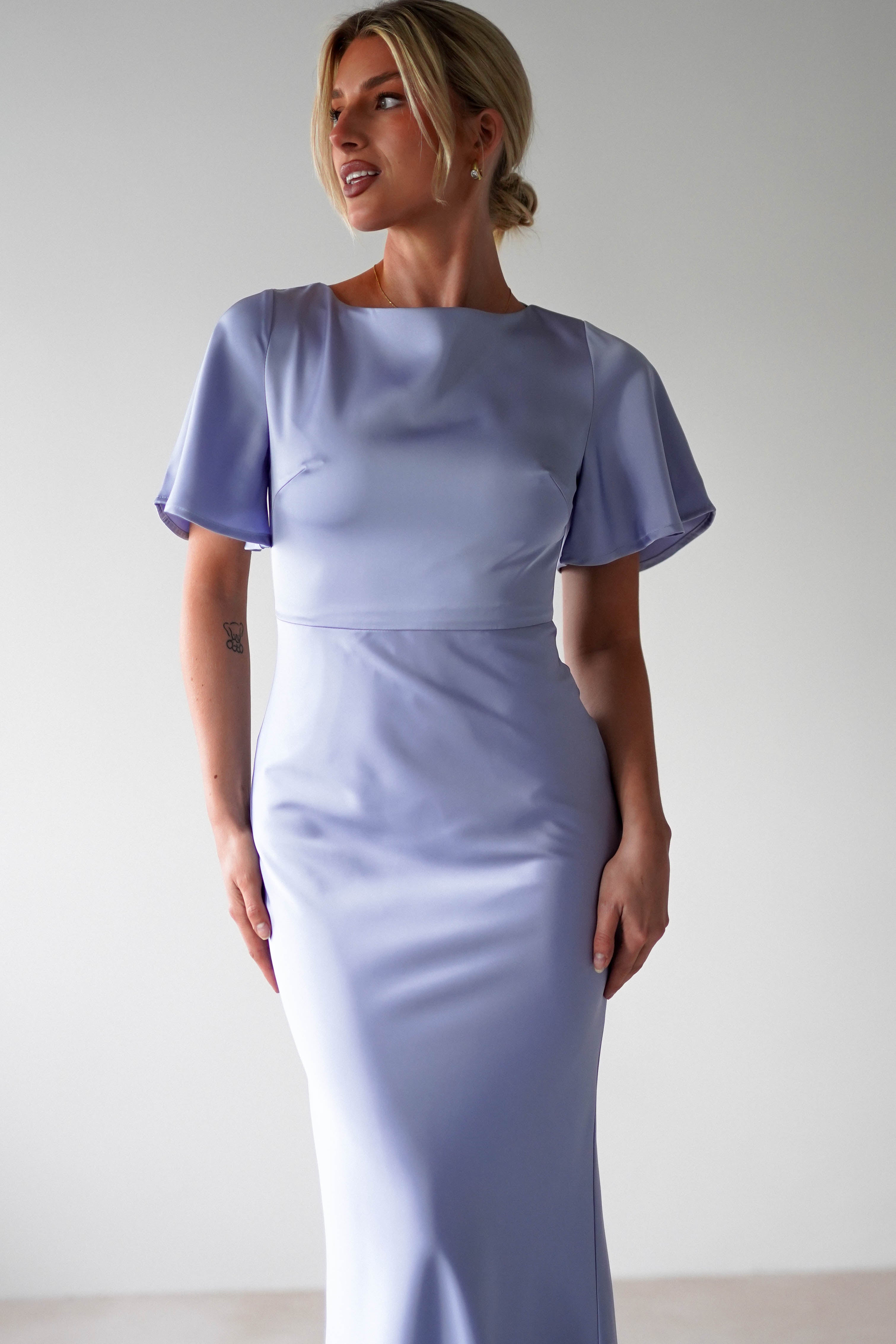 A woman wearing a light lavender satin maxi dress stands against a plain light background, looking to the side with her hair styled in a low updo.