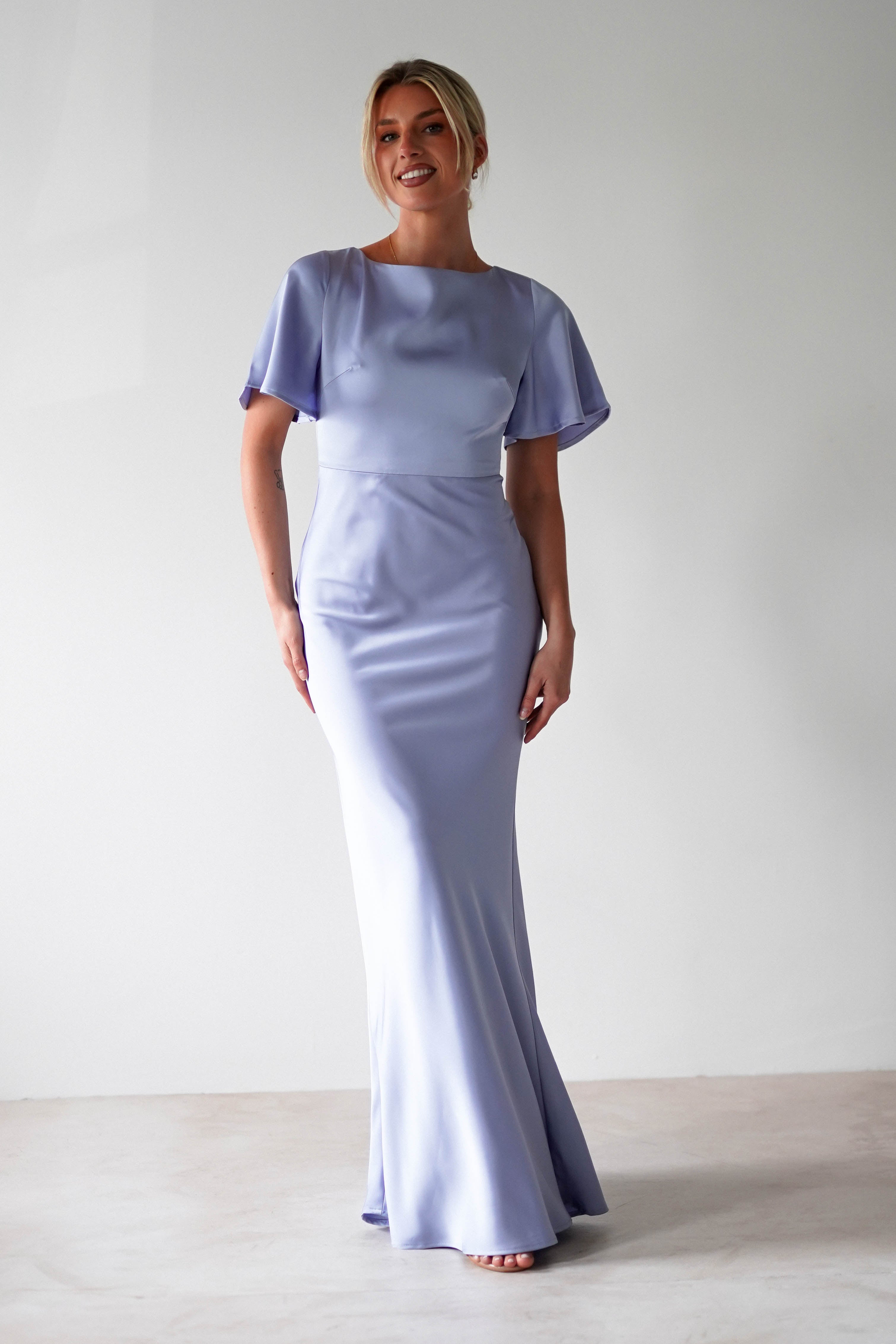 A woman in a light purple satin maxi dress with short flutter sleeves stands barefoot on a light floor, smiling in front of a plain white wall.