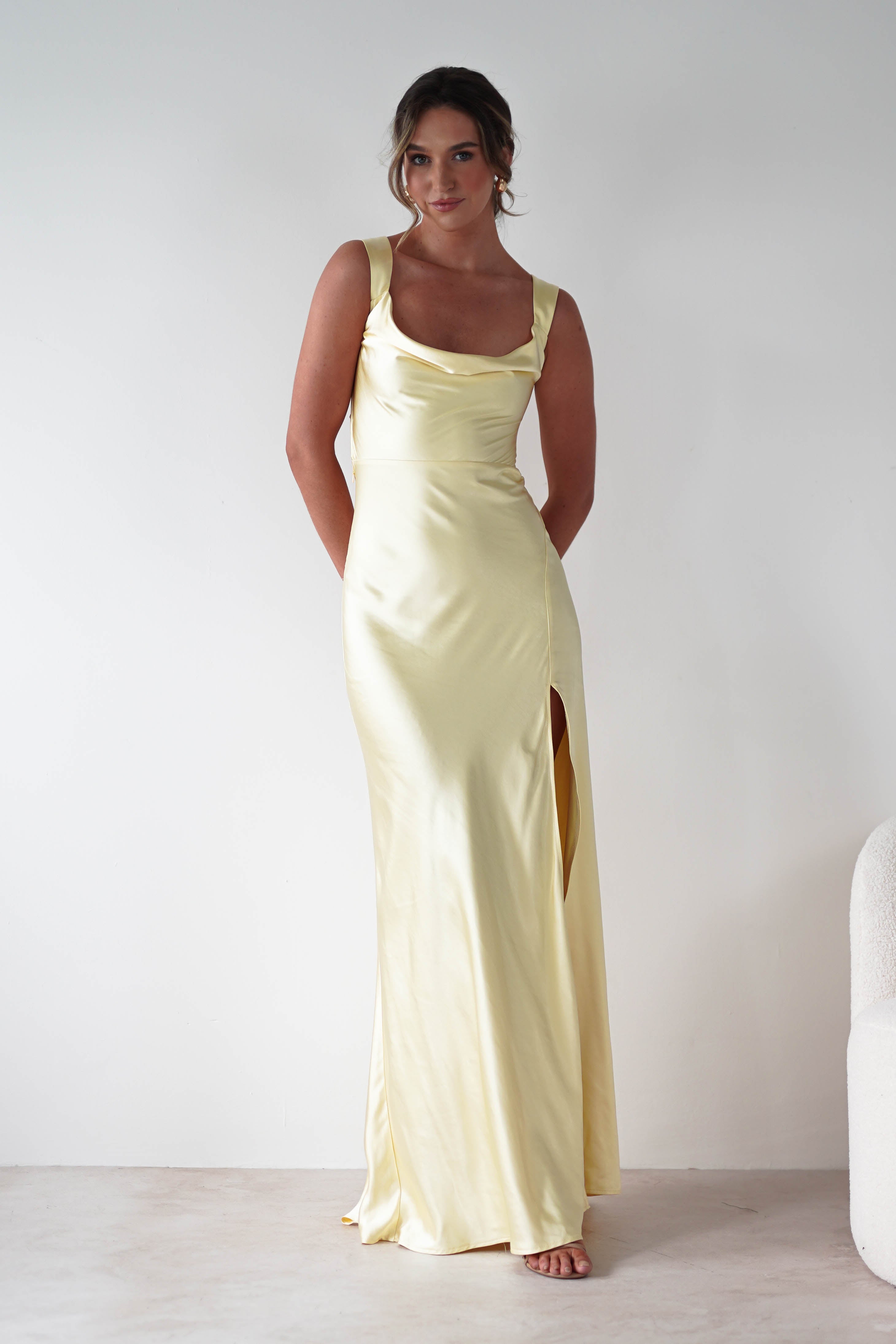 A woman stands barefoot against a white background, wearing a long yellow satin gown with a cowl neckline, side slit, and wide shoulder straps. She has her hands behind her back and her hair styled in a loose updo.