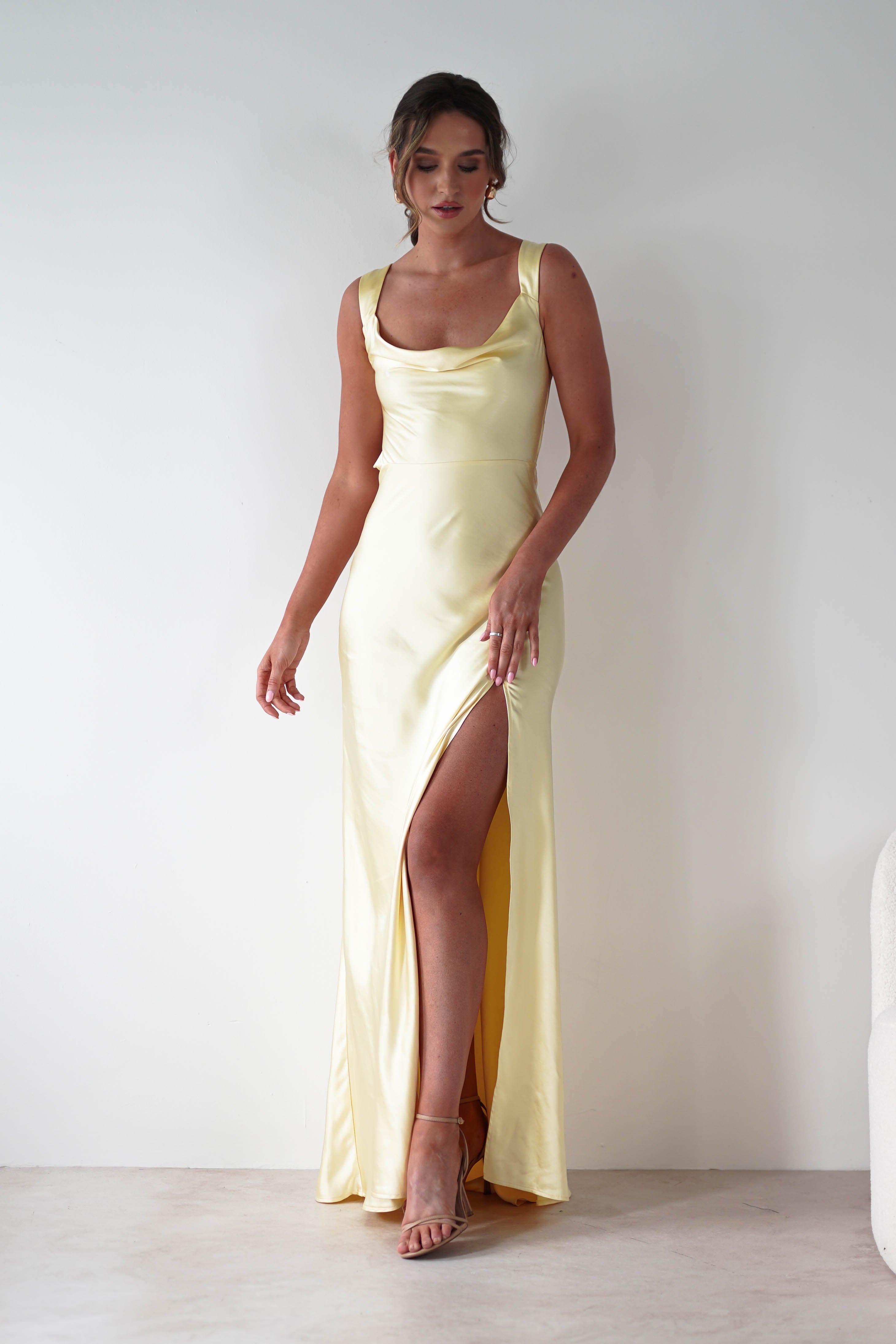 A woman stands against a white wall wearing a long yellow gown crafted from satin fabric, featuring a cowl neckline, thigh-high slit, spaghetti straps, styled hair, and high-heeled sandals.
