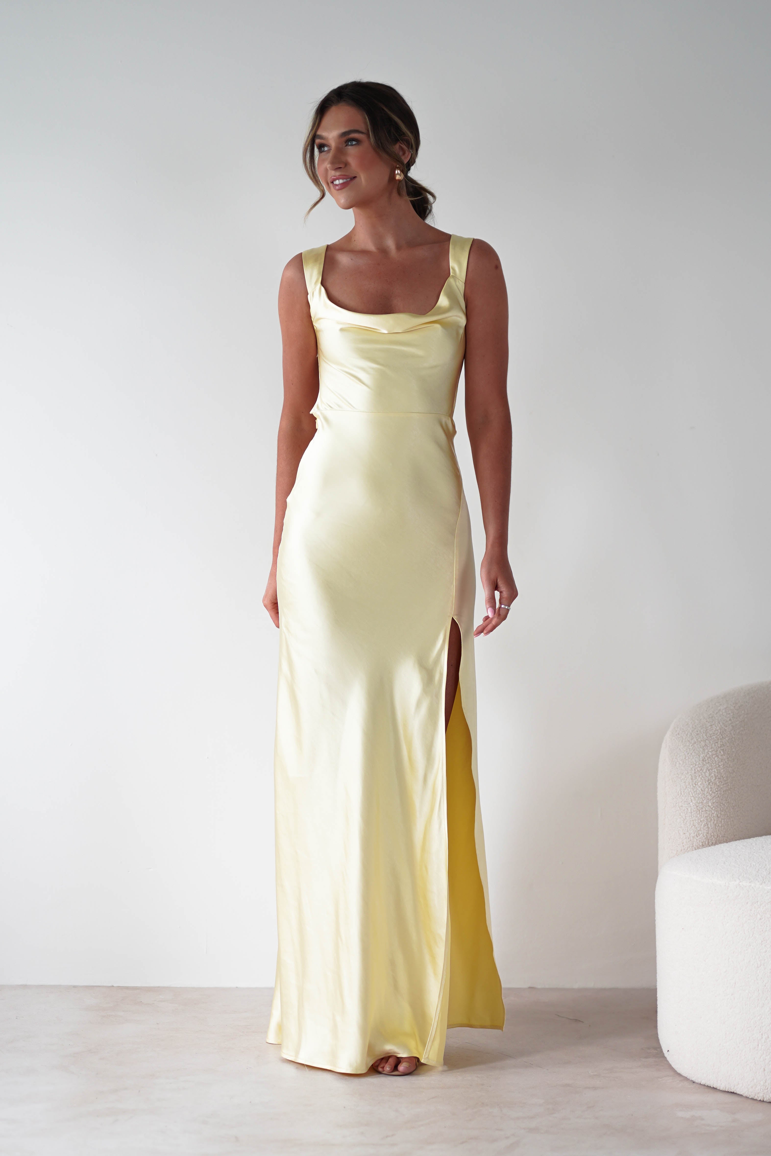 A woman wearing a long, pale yellow gown with a cowl neckline and thigh-high slit stands indoors against a plain white wall next to a light-colored upholstered chair. She is smiling and looking slightly to the side.