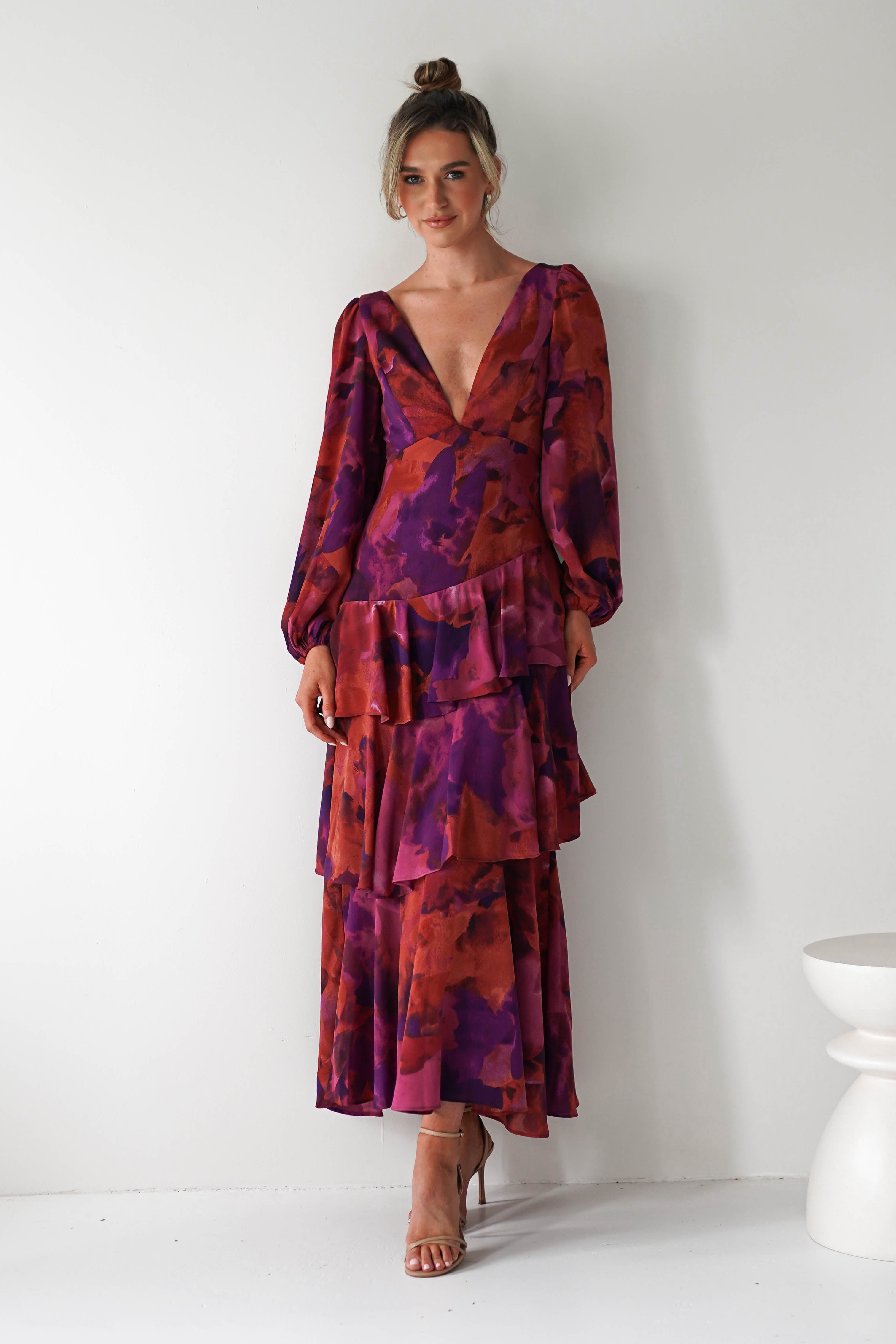 A woman stands against a white wall, wearing a flowing chiffon fabric maxi dress with a deep V neckline and long sleeves. The bold, abstract purple and red pattern makes it perfect for a stylish wedding guest look. She pairs it with nude heels and a top bun.