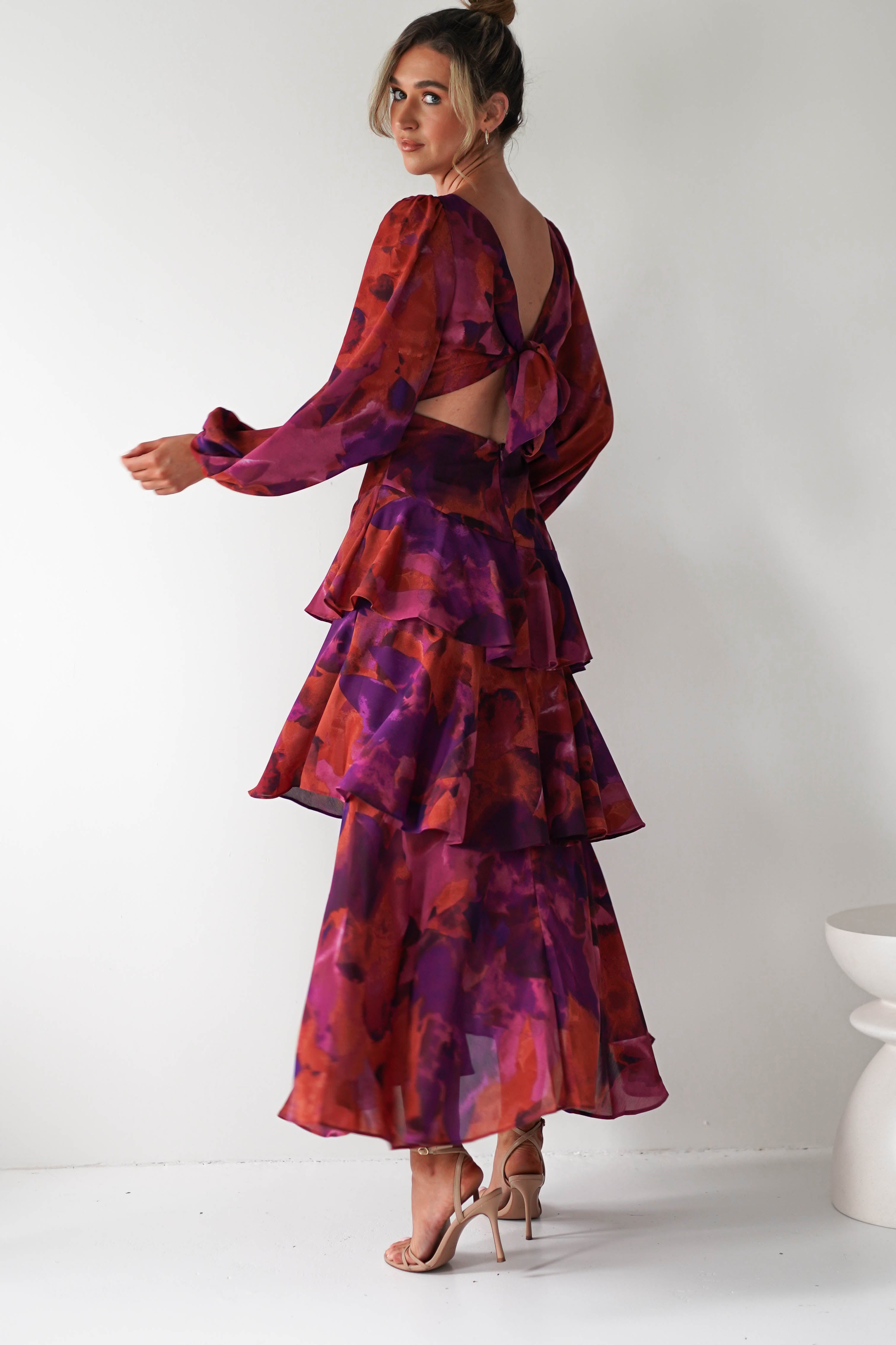 A woman poses in a purple and red floral ruffled maxi dress with an open back and tie detail, crafted from airy chiffon fabric—perfect for a wedding guest. She wears nude heels and stands against a white background, partially looking over her shoulder.