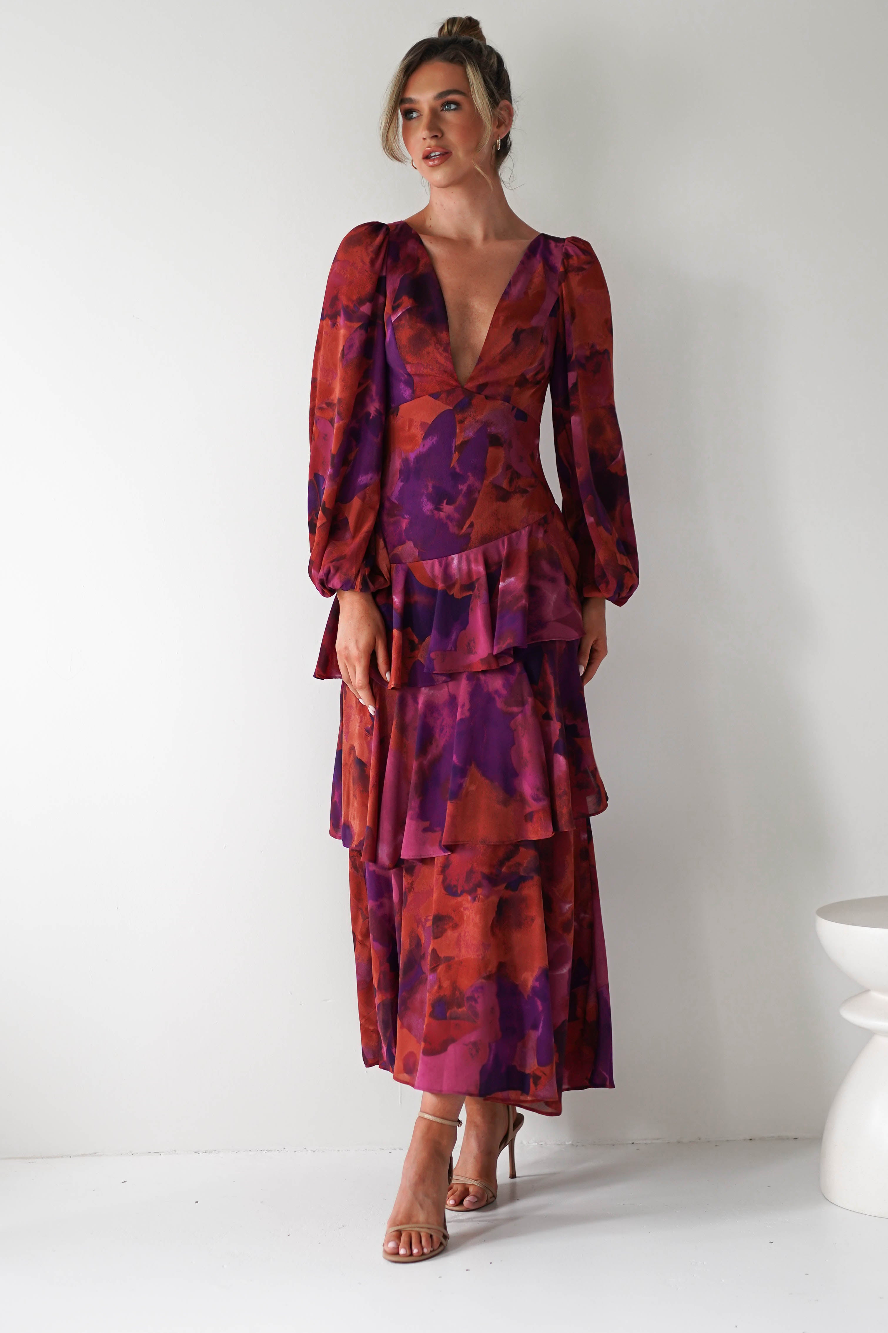 A woman wearing a chiffon fabric maxi dress with a deep V-neckline and long sleeves, featuring red, purple, and orange abstract florals—perfect for a stylish wedding guest—stands against a plain white background.