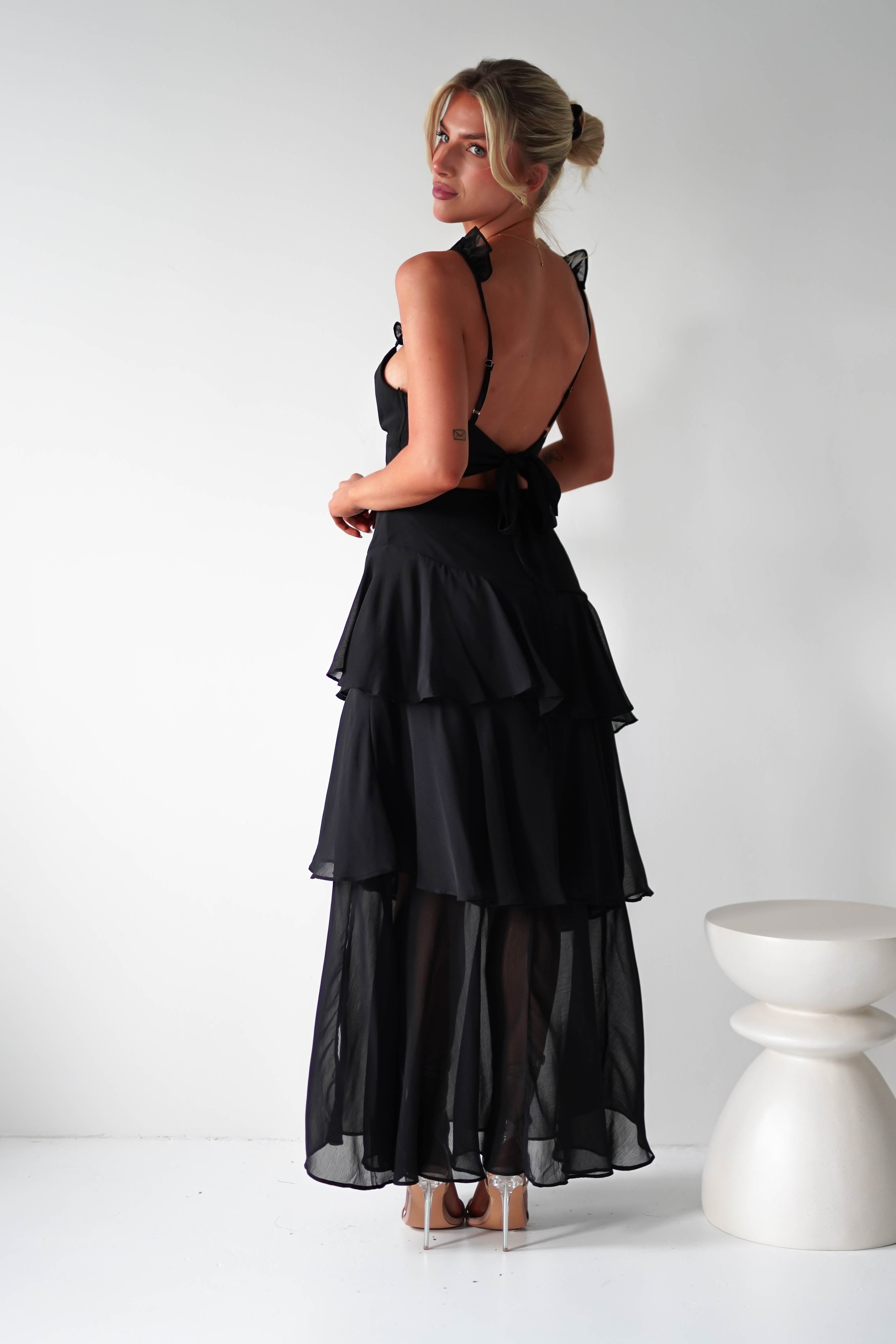 A person in a long, tiered ruffle dress with a low back and made of elegant chiffon fabric stands in a minimalist room. Their hair is in an updo, and they are wearing heels, perfect for a wedding guest. A white, sculptural stool is in the background.