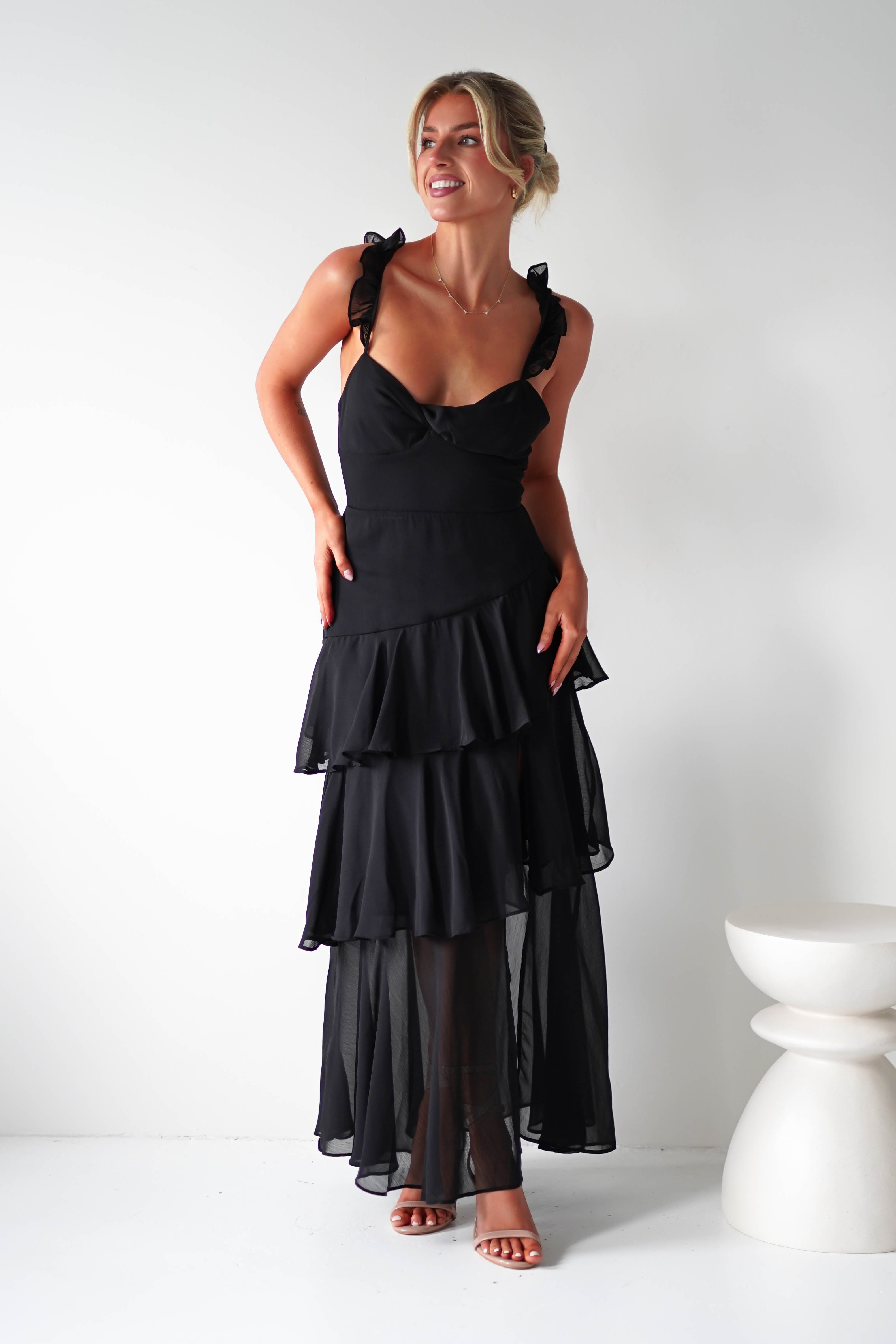 A woman in a sleeveless black ruffle dress, perfect for a wedding guest ensemble, stands against a white background, smiling. Her hair is elegantly tied up and she complements her look with high-heeled sandals. A small white round table is beside her.