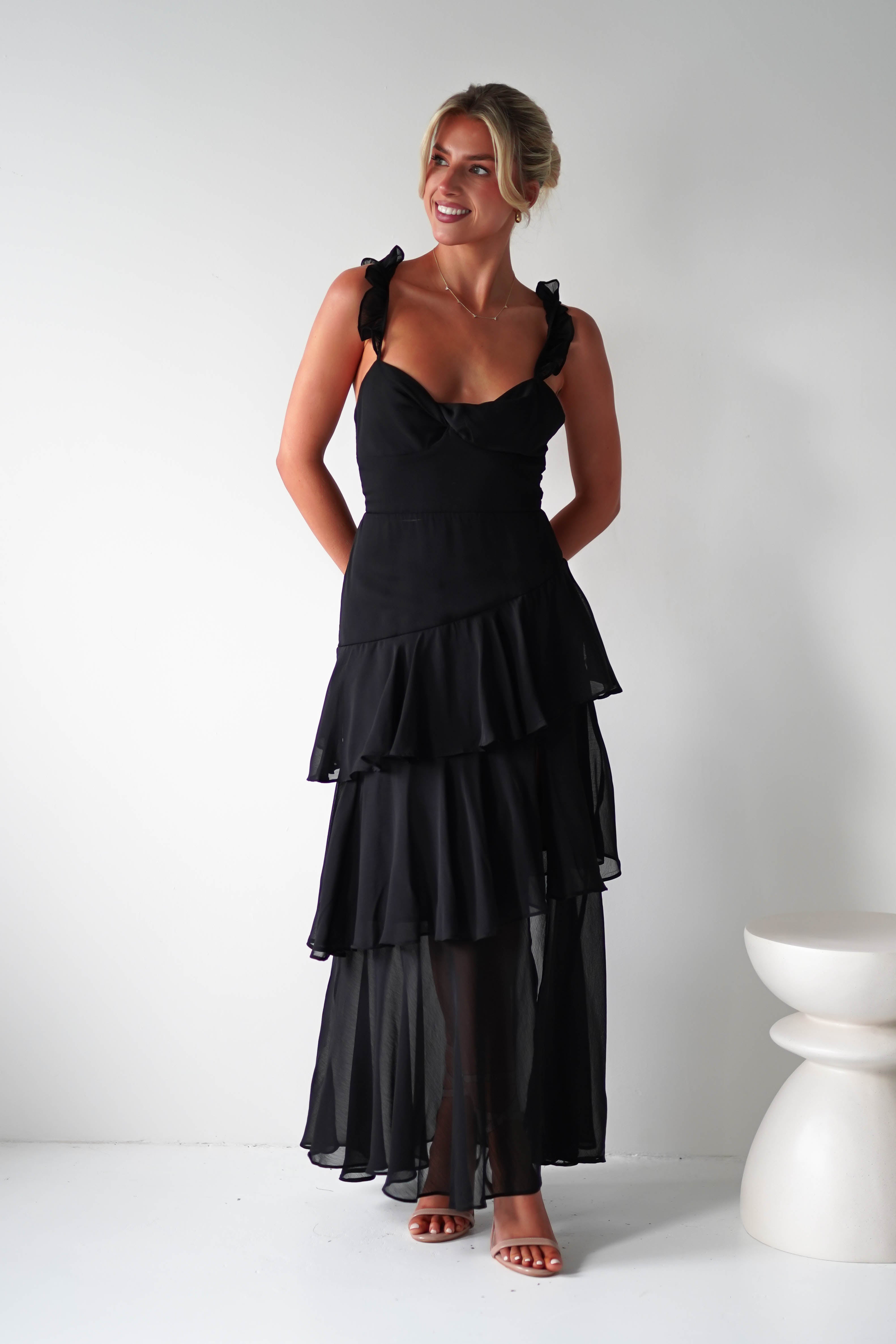 A person stands smiling in a black ruffle dress made of elegant chiffon fabric, perfect for a wedding guest, against a plain white background. Their hands are behind their back, and they wear brown sandals beside a small white table.