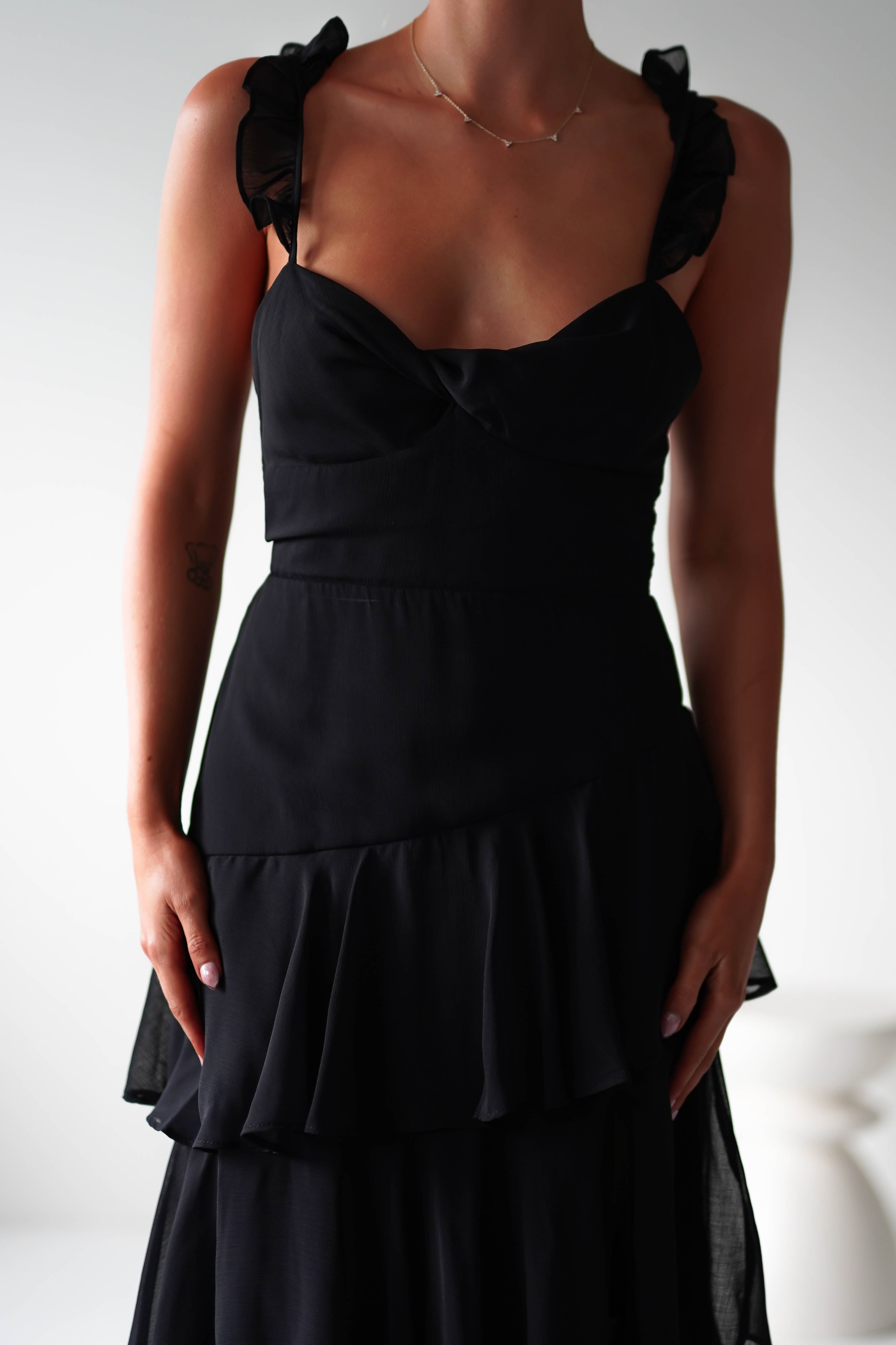 A person wearing a sleeveless black ruffle dress with chiffon layers and a sweetheart neckline stands in a well-lit, neutral space. Perfect attire for a wedding guest, only the upper body is visible.