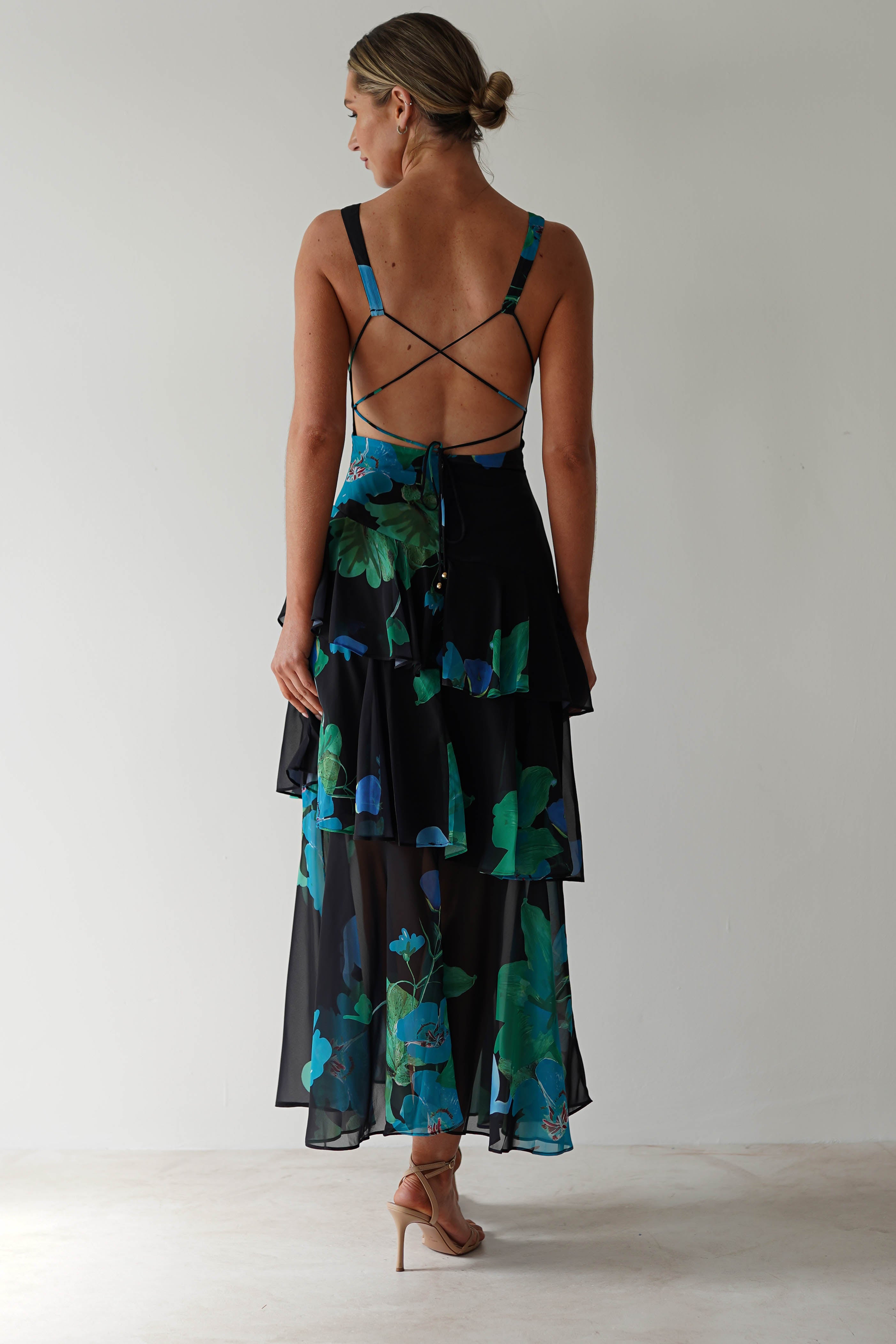 A person in a stunning black floral midi dress with an open back and tiered skirt stands against a plain background. The ensemble, perfect for wedding guest attire, features blue and green patterns on chiffon fabric. Their hair is in a bun, complemented by nude heeled sandals.