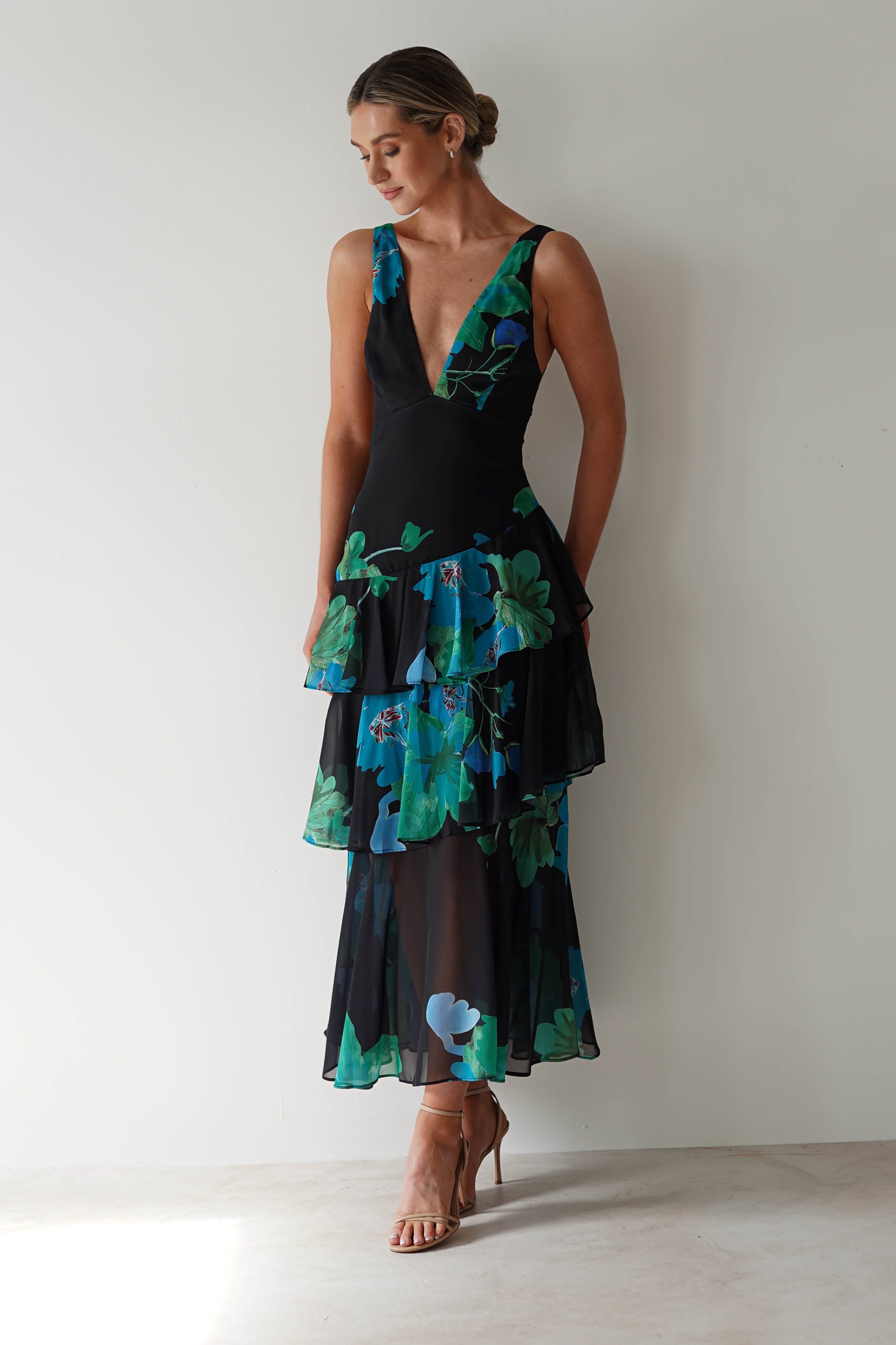 A woman in a sleeveless, V-neck black floral midi dress with layered patterns of blue and green stands against a light background. Perfect as a wedding guest outfit, she pairs it with nude high-heeled sandals and styles her hair in a low bun.