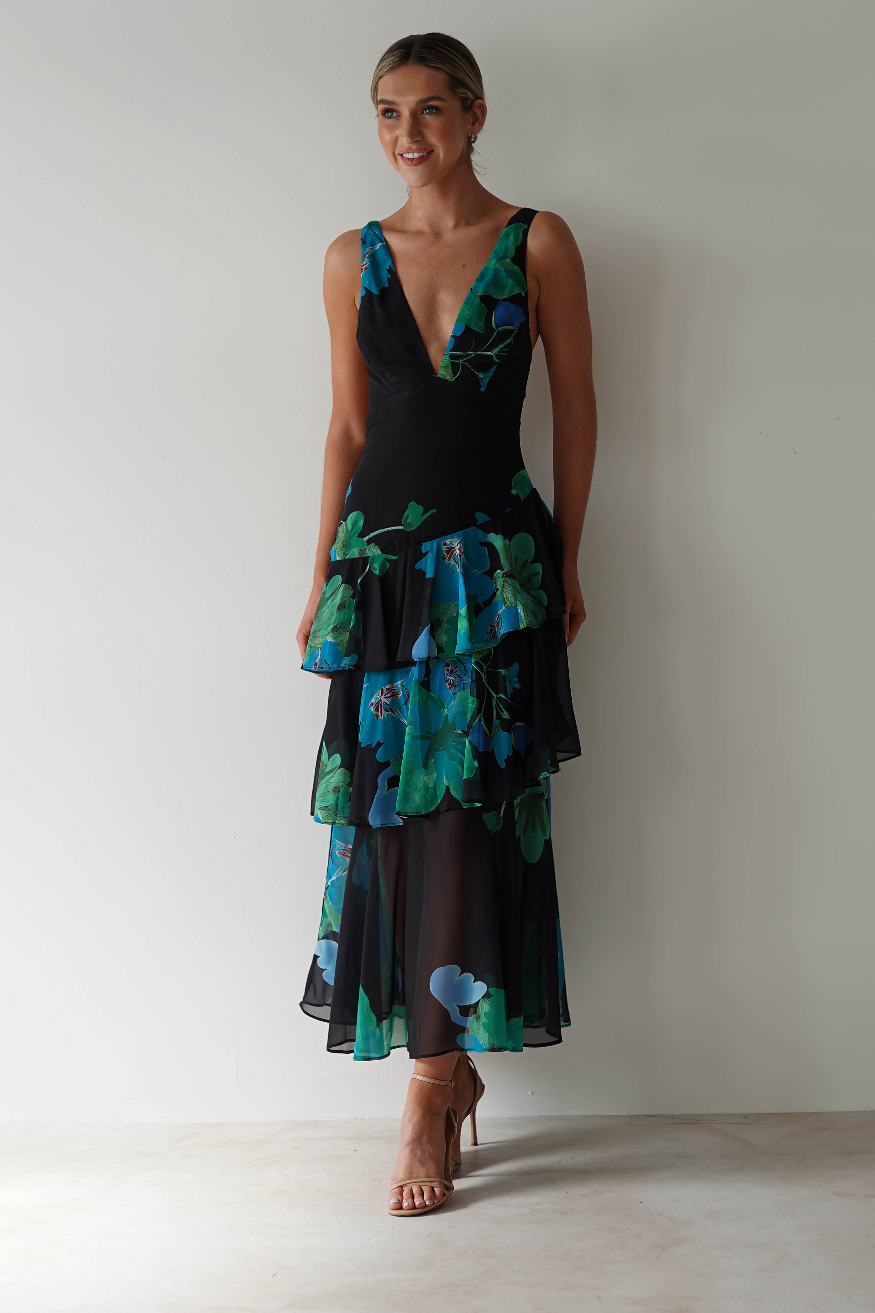 A person stands against a plain wall in a sleeveless, black floral midi dress with vibrant green and blue patterns. The chiffon fabric adds an elegant touch as they smile, hair tied back, and wear strappy heels—perfect wedding guest attire.