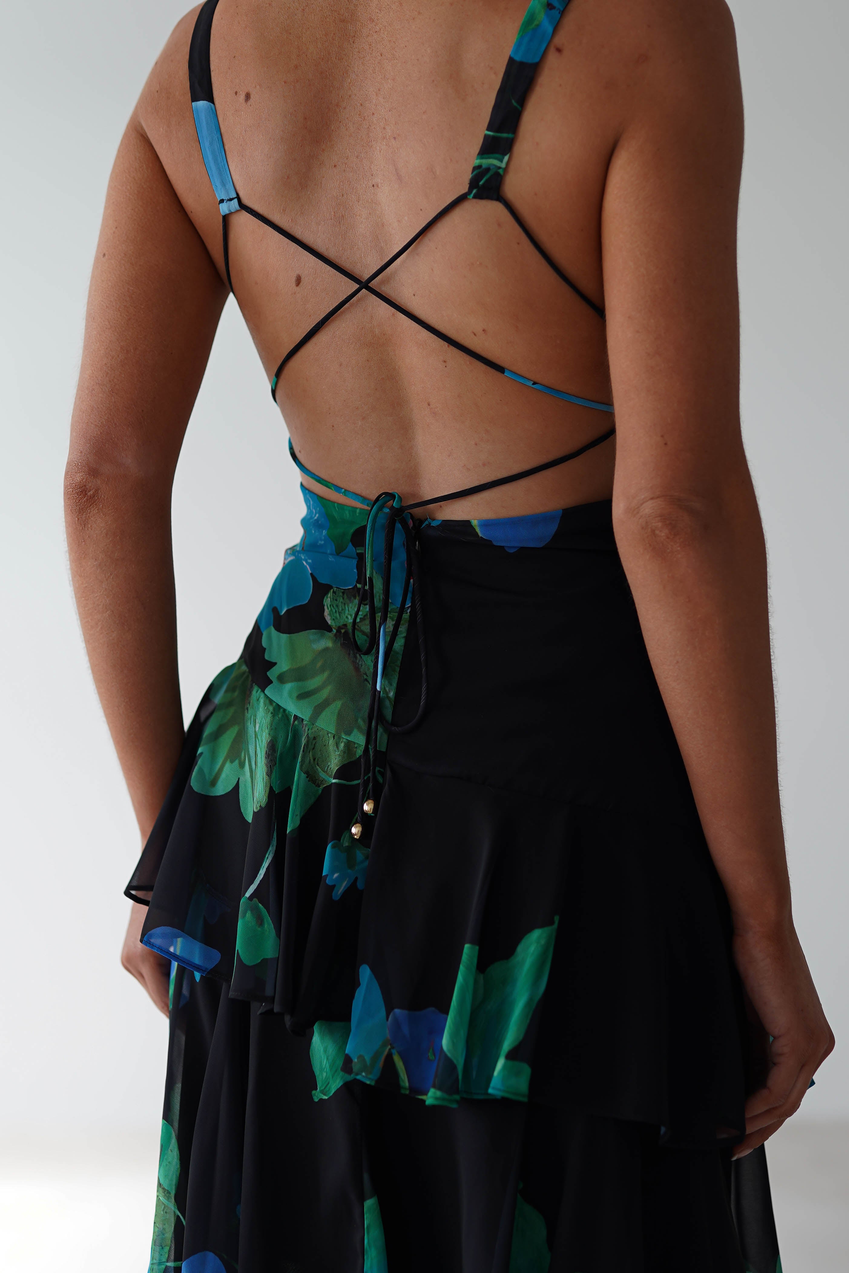 A person wearing a chic black floral midi dress stands with their back to the camera. The sleeveless outfit, perfect as a wedding guest outfit, features thin crisscross straps and a tie at the back, showcasing a floral pattern in shades of blue and green.
