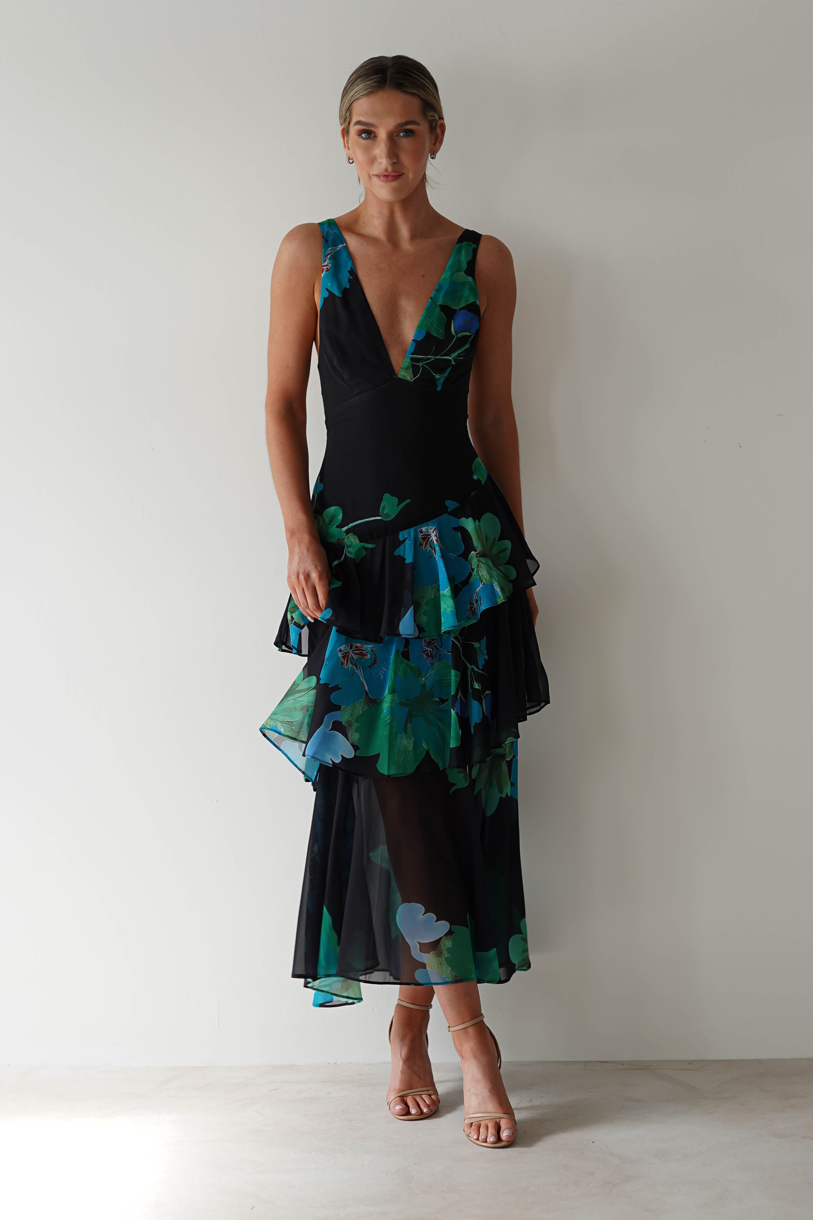 A person stands against a plain background, wearing a black floral midi dress with vibrant blue and green patterns. The chiffon dress features layered ruffles and a deep V-neck, ideal as a wedding guest outfit. Nude heels complete the ensemble elegantly.