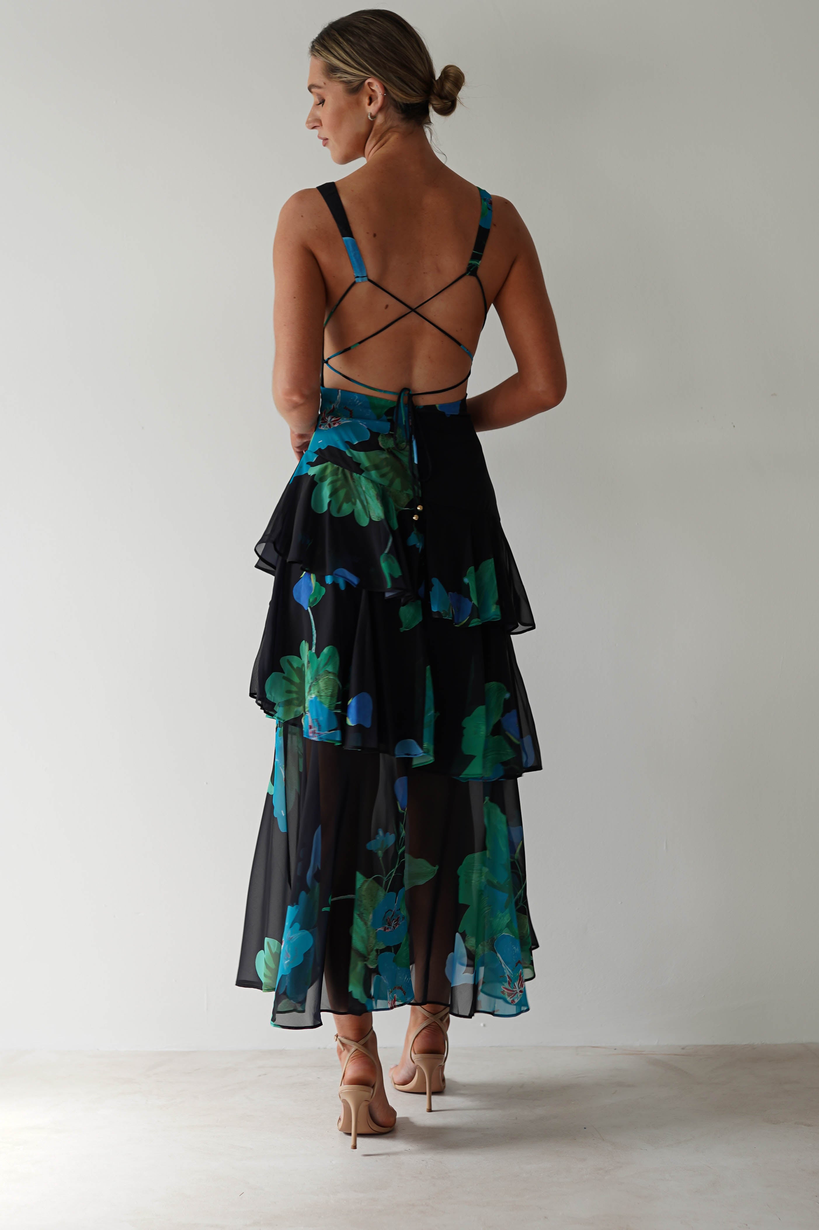 A person with hair up in a bun is wearing a black floral midi dress featuring green and blue patterns on chiffon fabric. The layered skirt and crisscross straps on the open back add elegance as they stand against a plain white background, perfect for a stylish wedding guest.