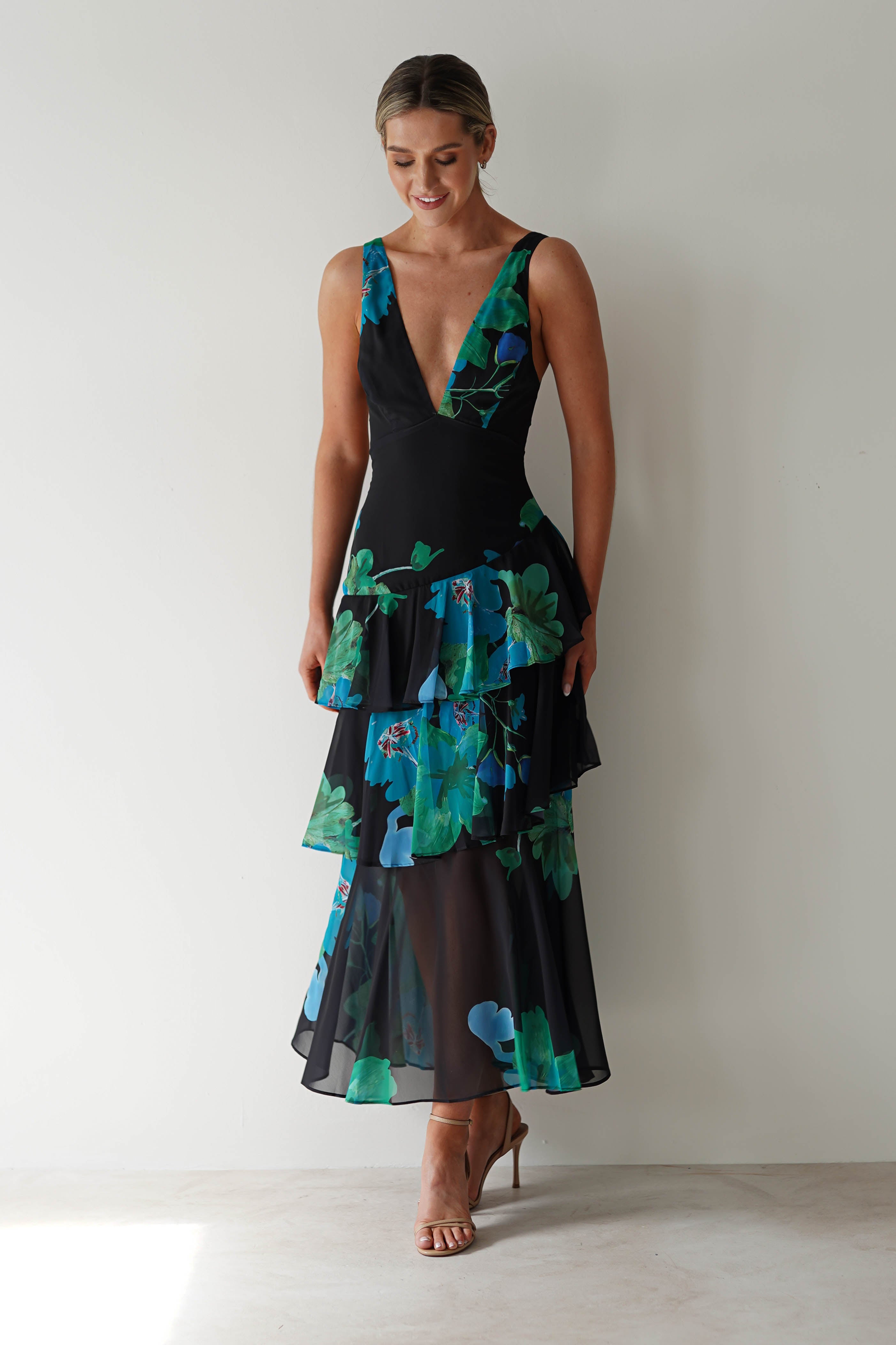 A woman in a sleeveless black and blue floral chiffon dress with a deep V-neck and tiered skirt stands against a plain wall, smiling softly with her hands at her sides. Perfect as a wedding guest outfit, she wears high heels and has her hair styled up.