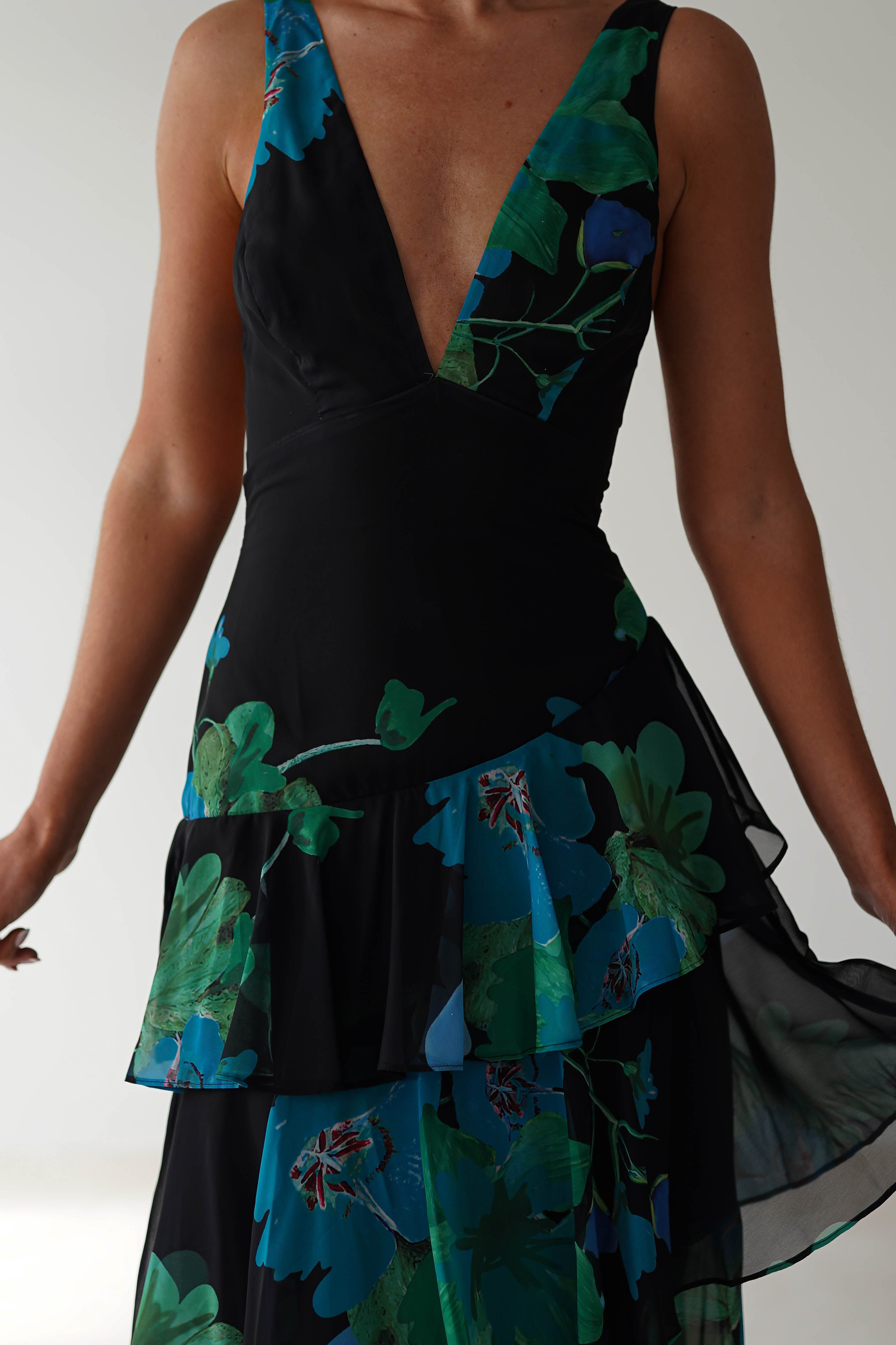 A person wearing a chic black floral midi dress with a deep V-neckline and layered patterns in blue and green hues. This chiffon dress, ideal as a wedding guest outfit, boasts a flowing design with tiered ruffles against a simple off-white backdrop.