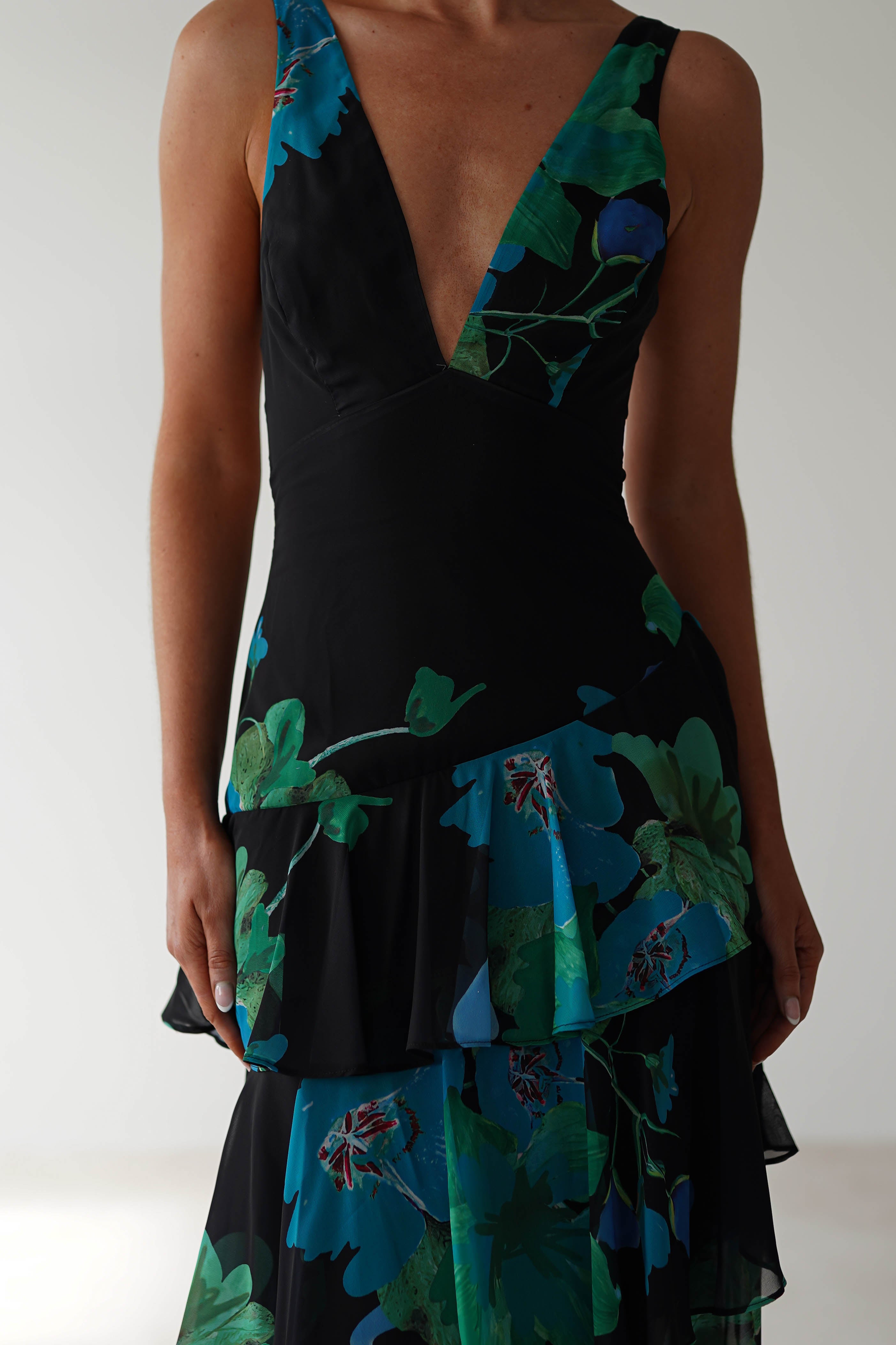 A person elegantly wears a black floral midi dress, showcasing a deep V-neckline. The chiffon fabric is adorned with green and blue patterns and layered ruffles, perfect for a wedding guest. The simple white background highlights the beauty of the dress.