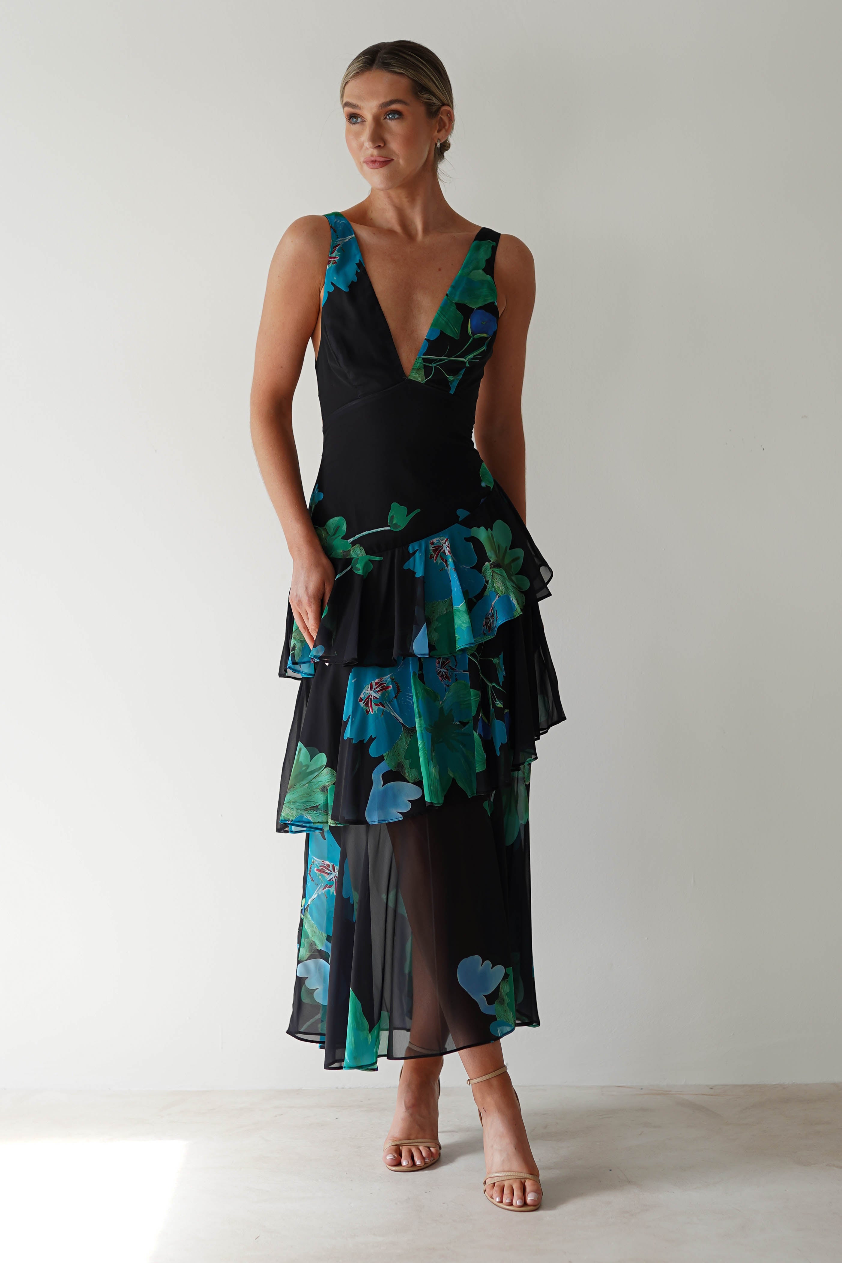 A person wearing a black floral midi dress crafted from chiffon fabric, adorned with layers of blue and green patterns. The dress features a deep V-neckline and a tiered skirt. They stand against a plain background, looking slightly to the side, perfect for wedding guest attire.