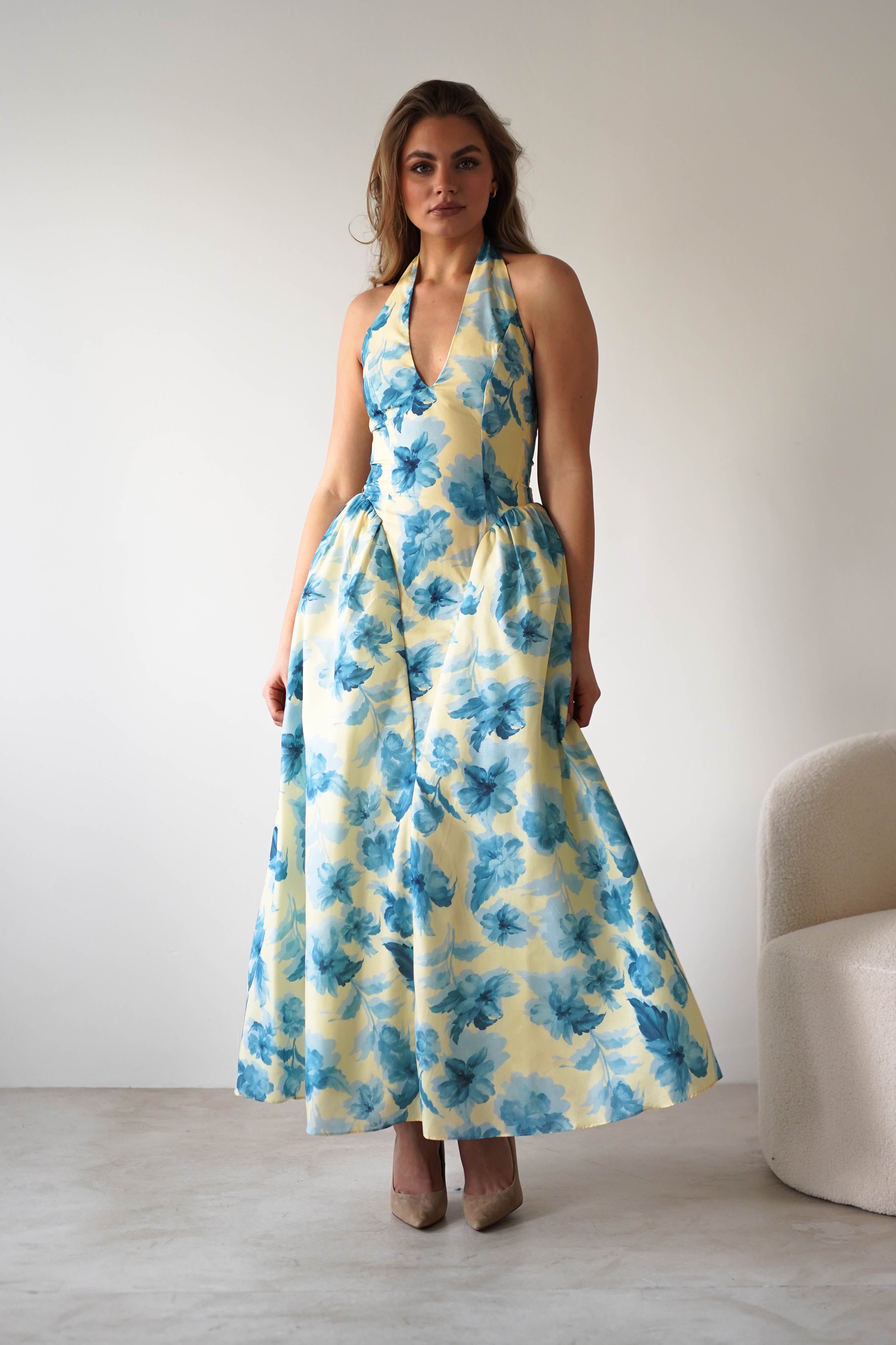 A woman stands indoors in a summer floral midaxi dress with a halter neckline and blue flowers on light yellow. She pairs beige heels, perfect as a chic wedding guest look, and poses near a white chair against a plain wall.