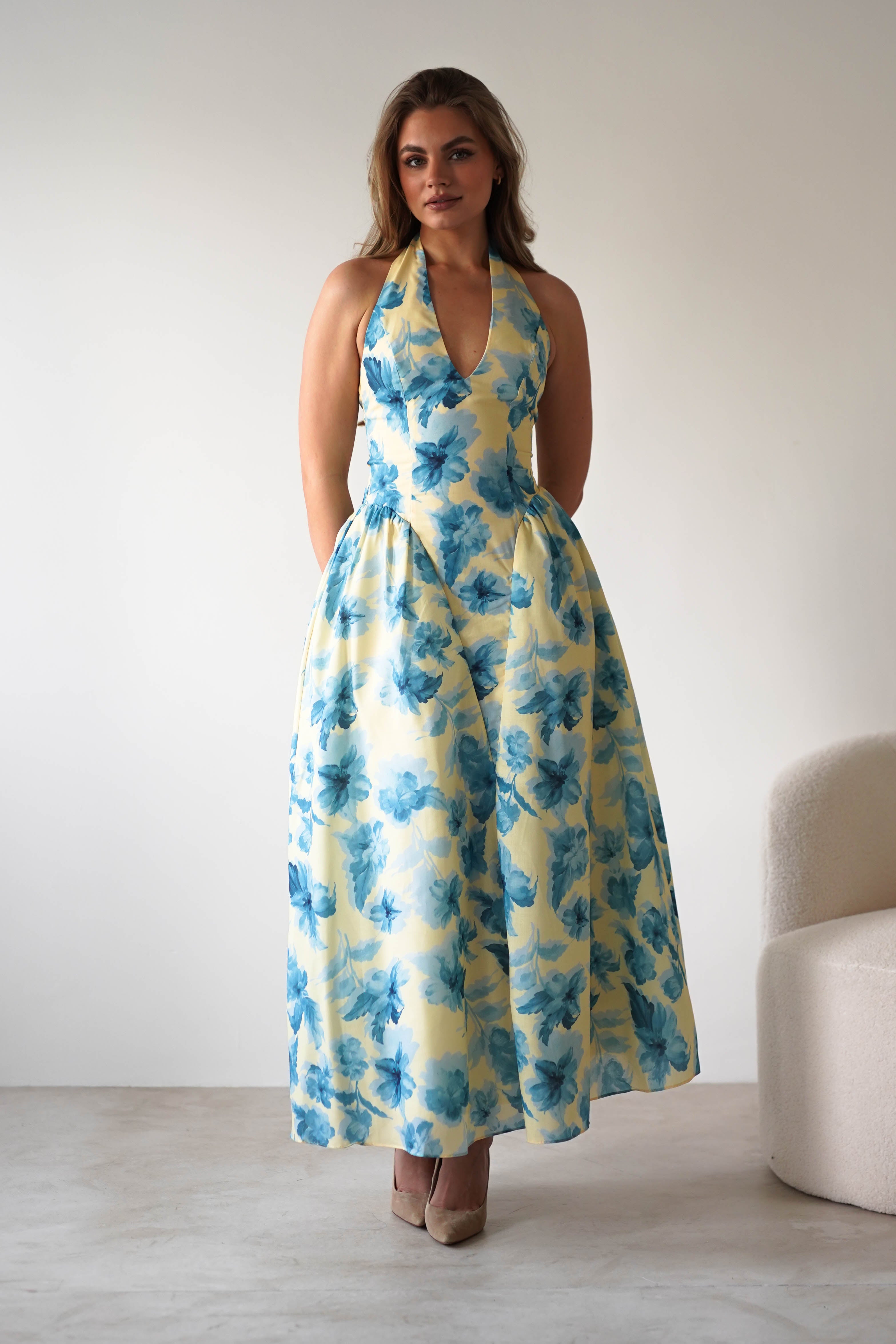A woman stands indoors wearing a summer floral midaxi dress with a halter-neck and ankle length. Her long hair cascades down as she pairs the look with beige high heels—an elegant choice for a wedding guest. The backdrop features a minimal white wall and chair.