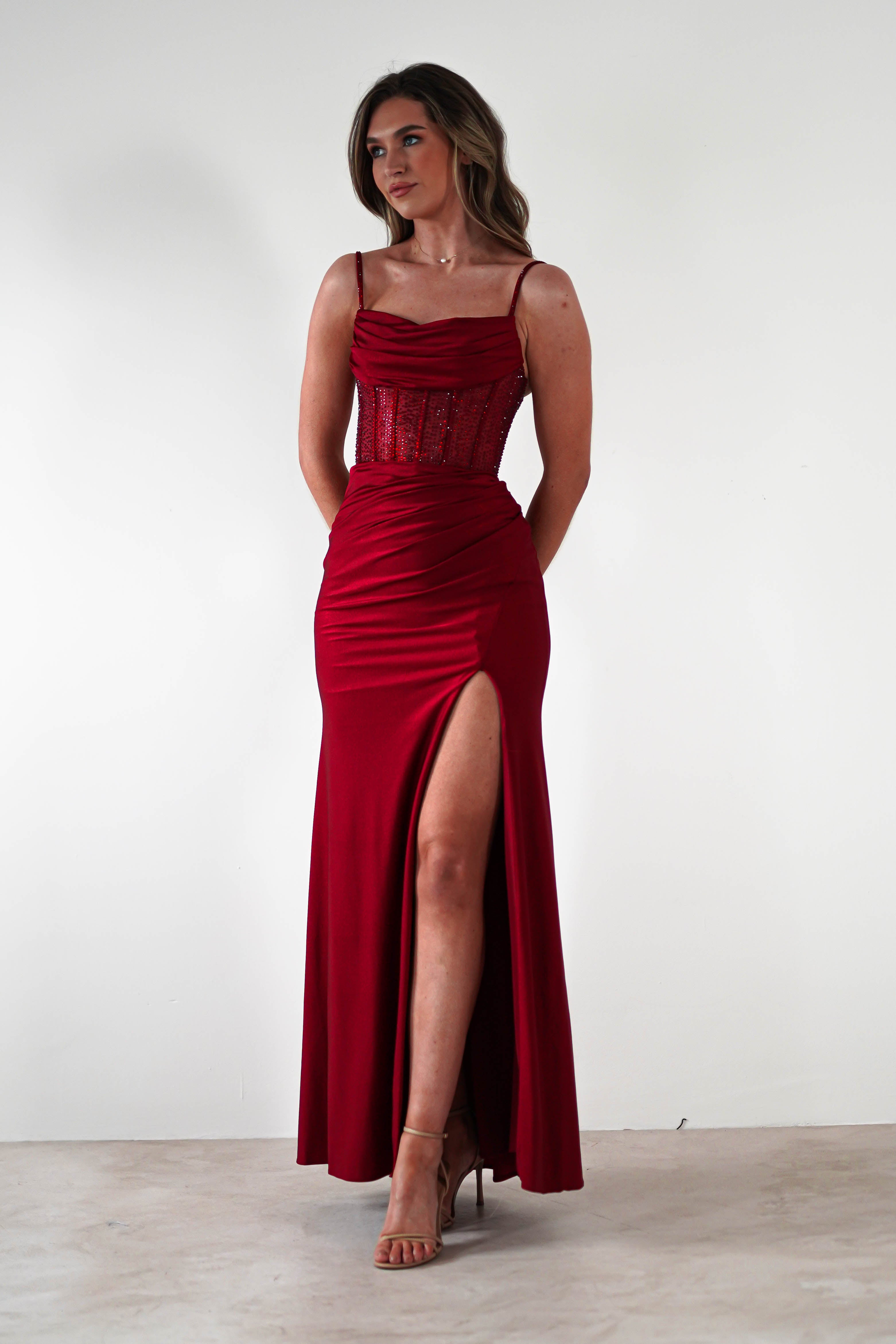 A woman in a sleeveless, floor-length red fitted gown with a rhinestone bodice and thigh-high slit stands against a plain white background, looking slightly to the side. Perfect for formal events, she completes the look with beige high-heeled sandals.