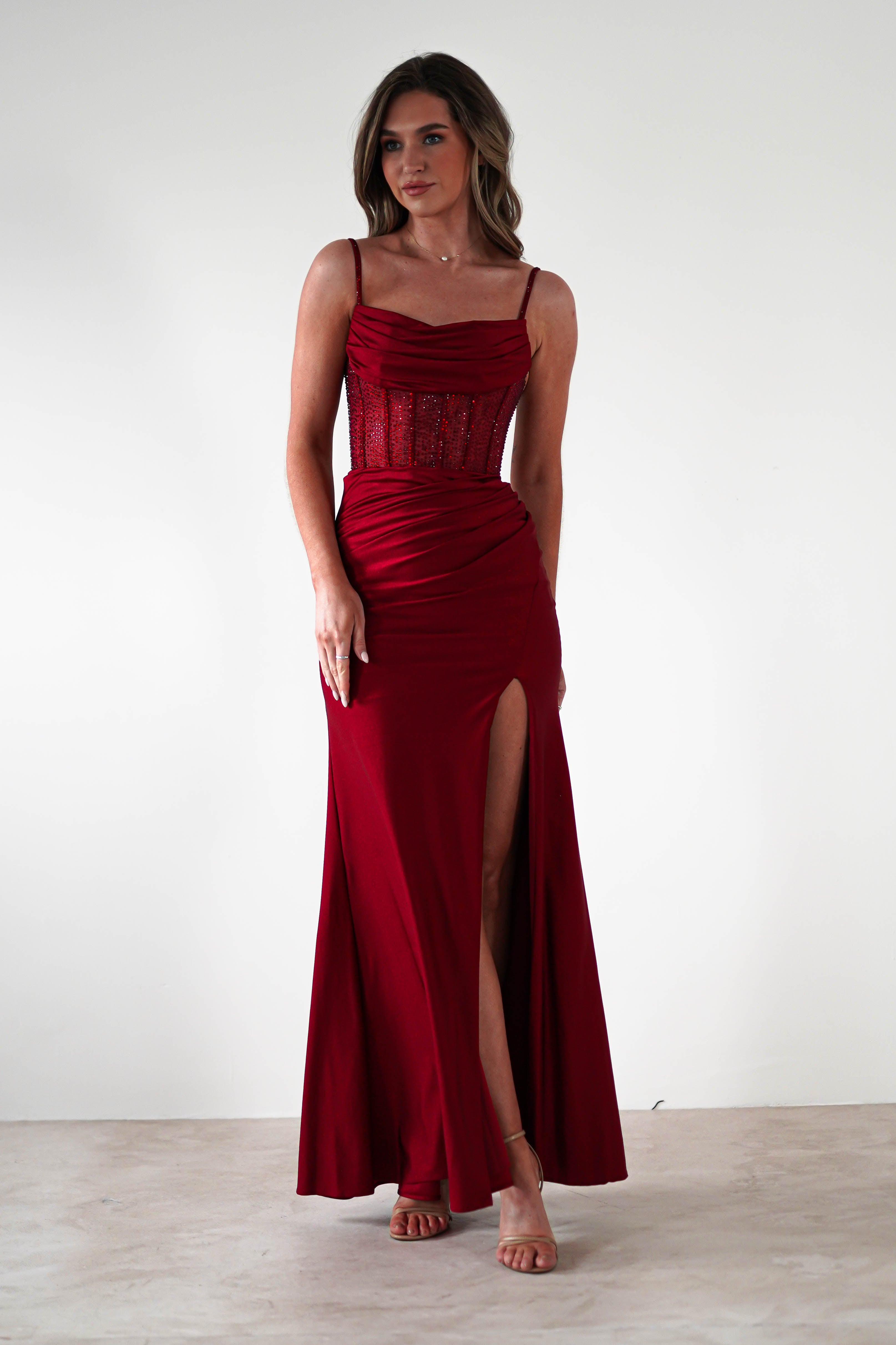 A woman wearing a sleeveless, floor-length red fitted gown with a rhinestone bodice, ruched details, and a high slit, standing against a plain white background—perfect for formal events.