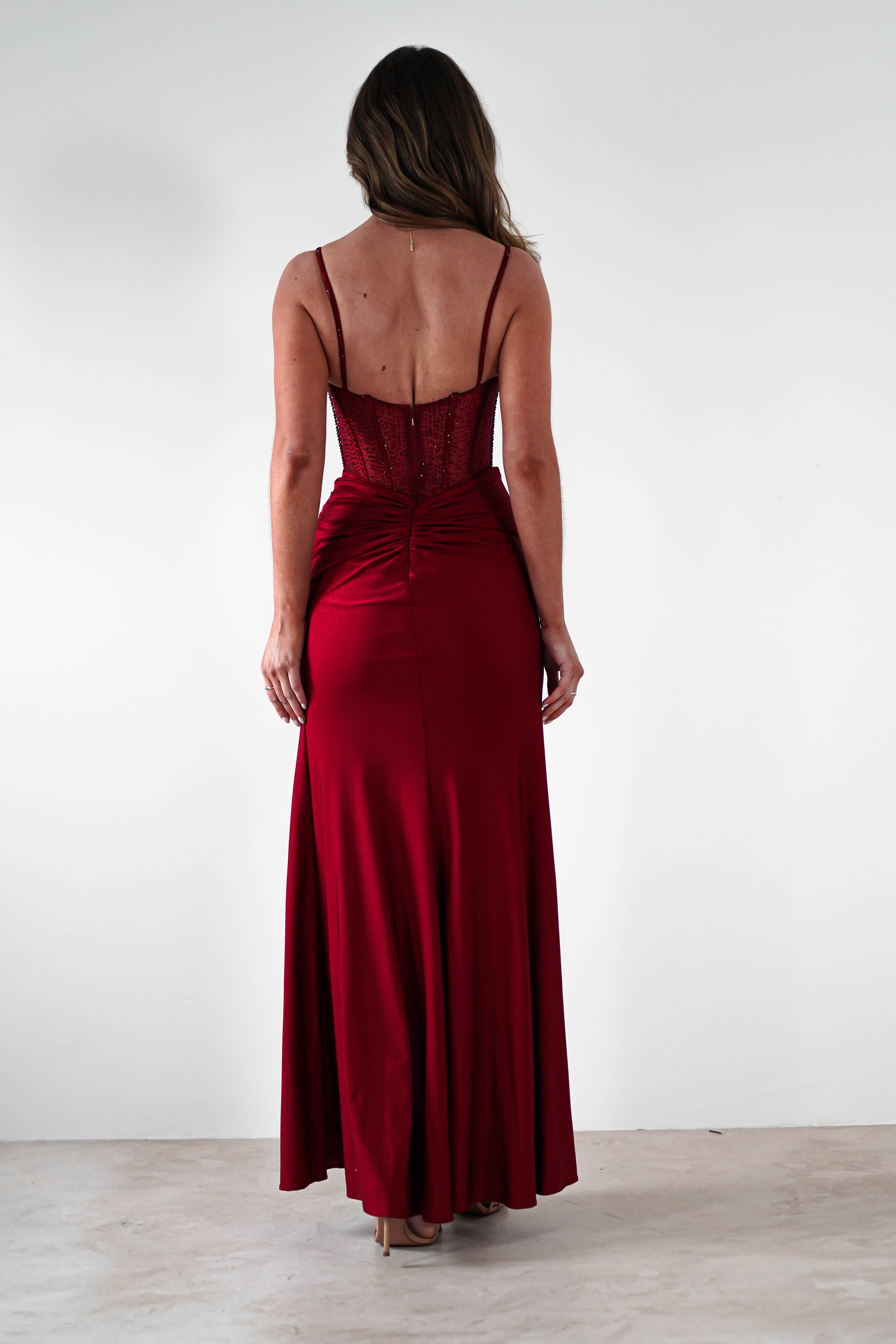 A woman with long hair stands facing away in a fitted red gown featuring a rhinestone bodice and thin straps, its floor-length silhouette and ruched back perfectly suited for formal events, set against a plain white background.