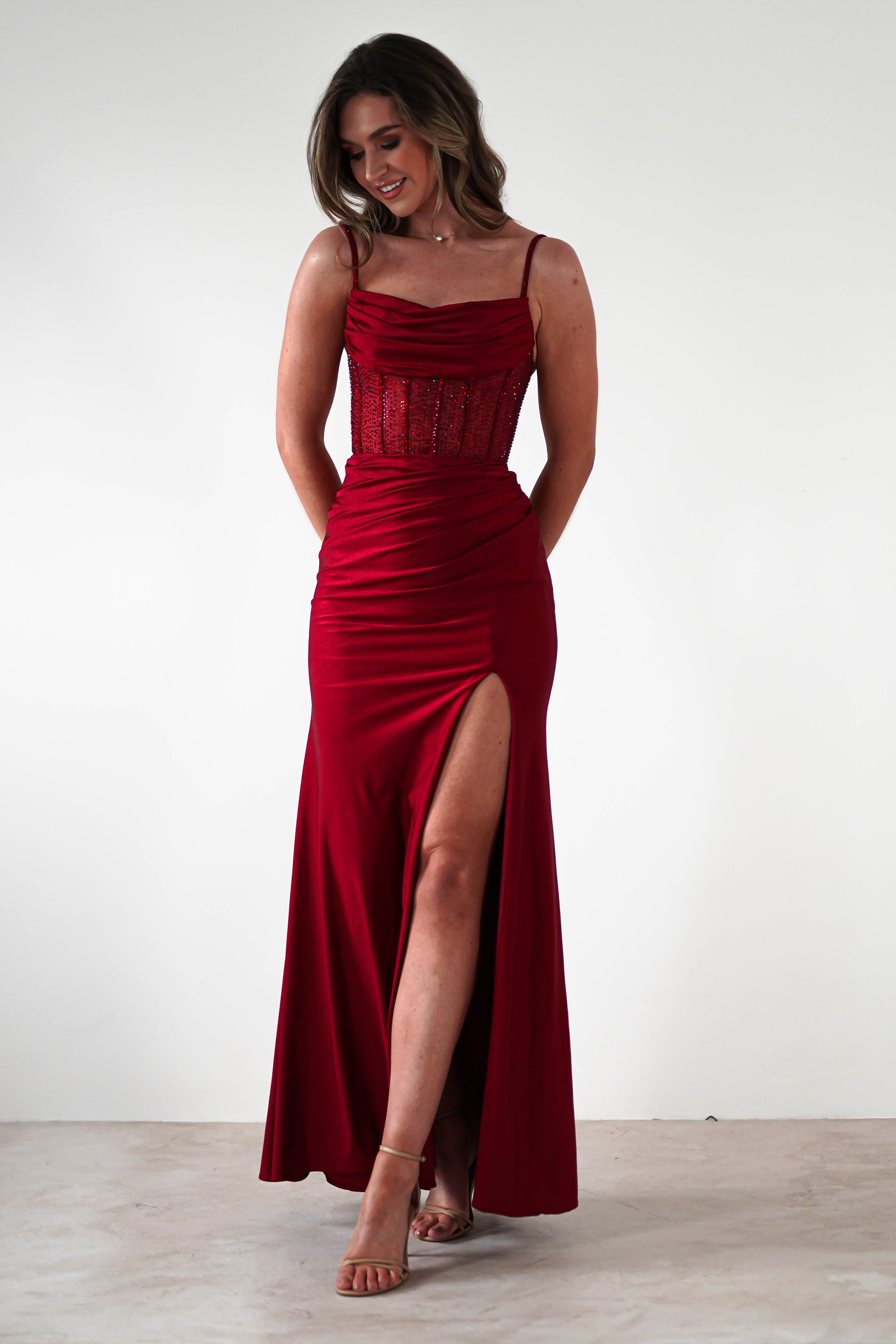 A woman in a sleeveless, floor-length fitted gown with a rhinestone bodice and thigh-high slit stands against a plain white background, smiling and looking down, with her hands behind her back—perfect for formal events. She wears open-toe heels.