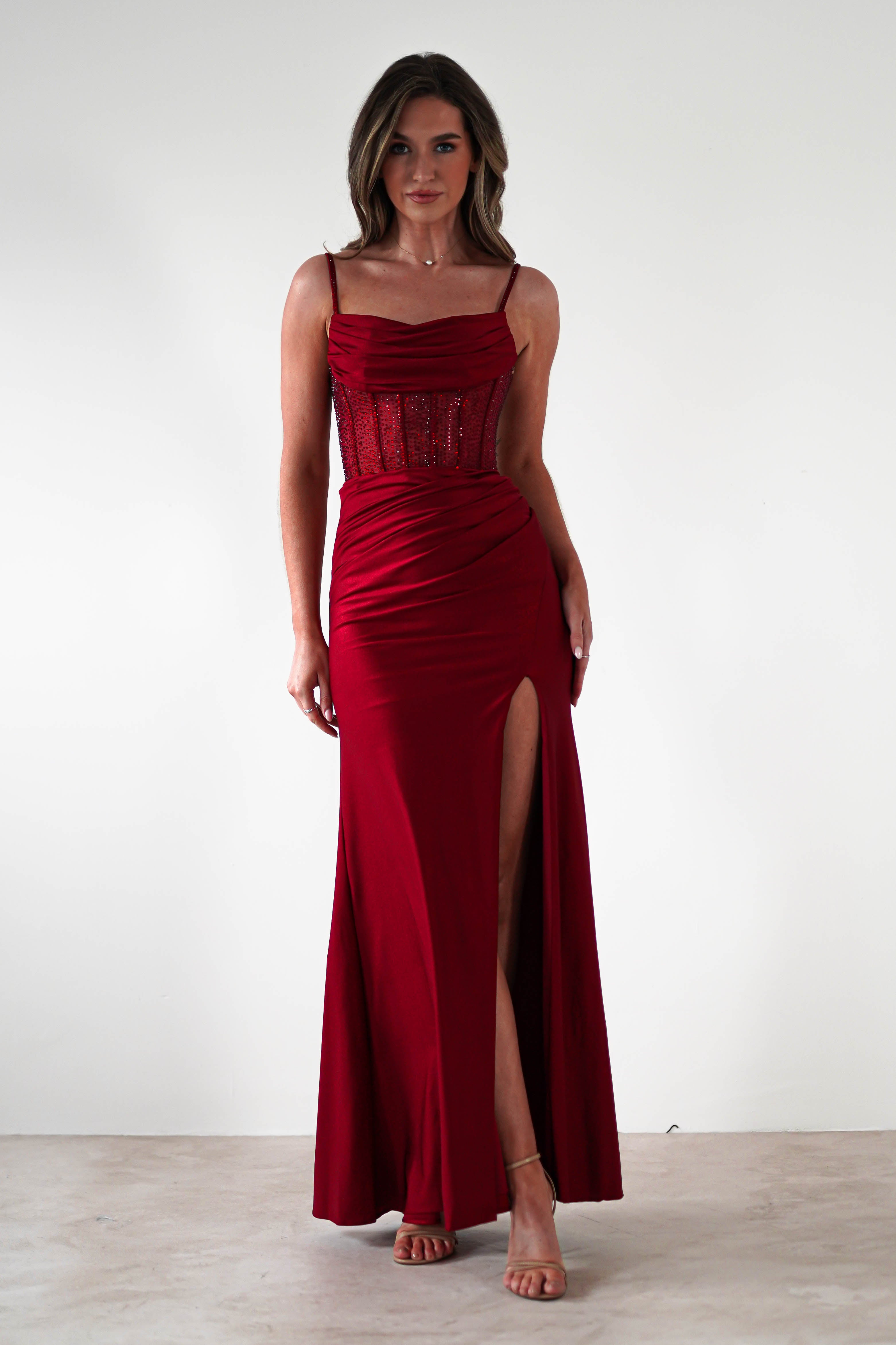 A woman stands against a plain white background wearing a fitted, floor-length red gown with a thigh-high slit and rhinestone bodice—perfect for formal events. She has shoulder-length hair and is wearing strappy heels.