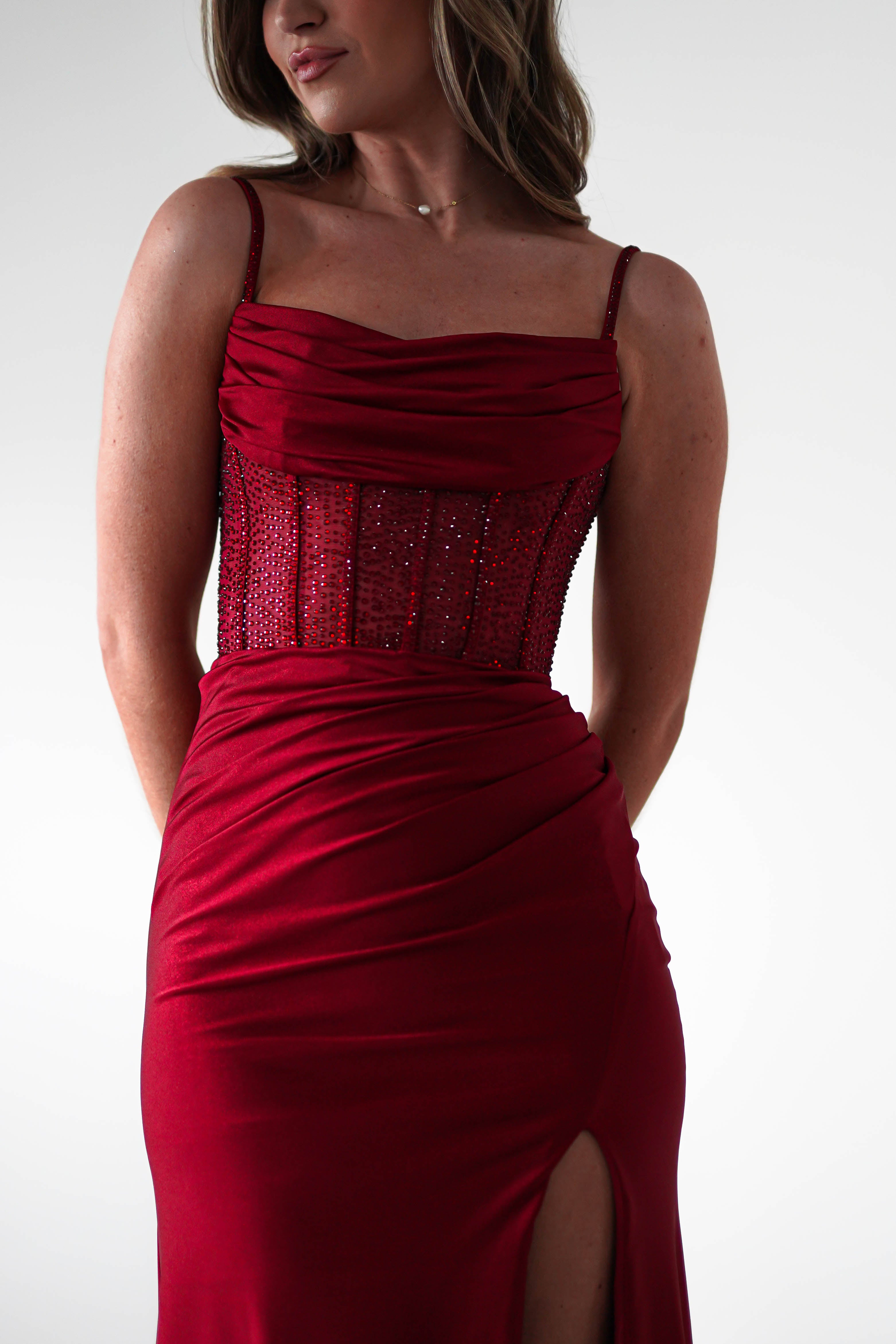 A woman in a sleeveless, fitted gown with a rhinestone bodice, draped skirt, and thigh-high slit stands against a plain light background. Her head is slightly turned to the side, perfect attire for formal events.