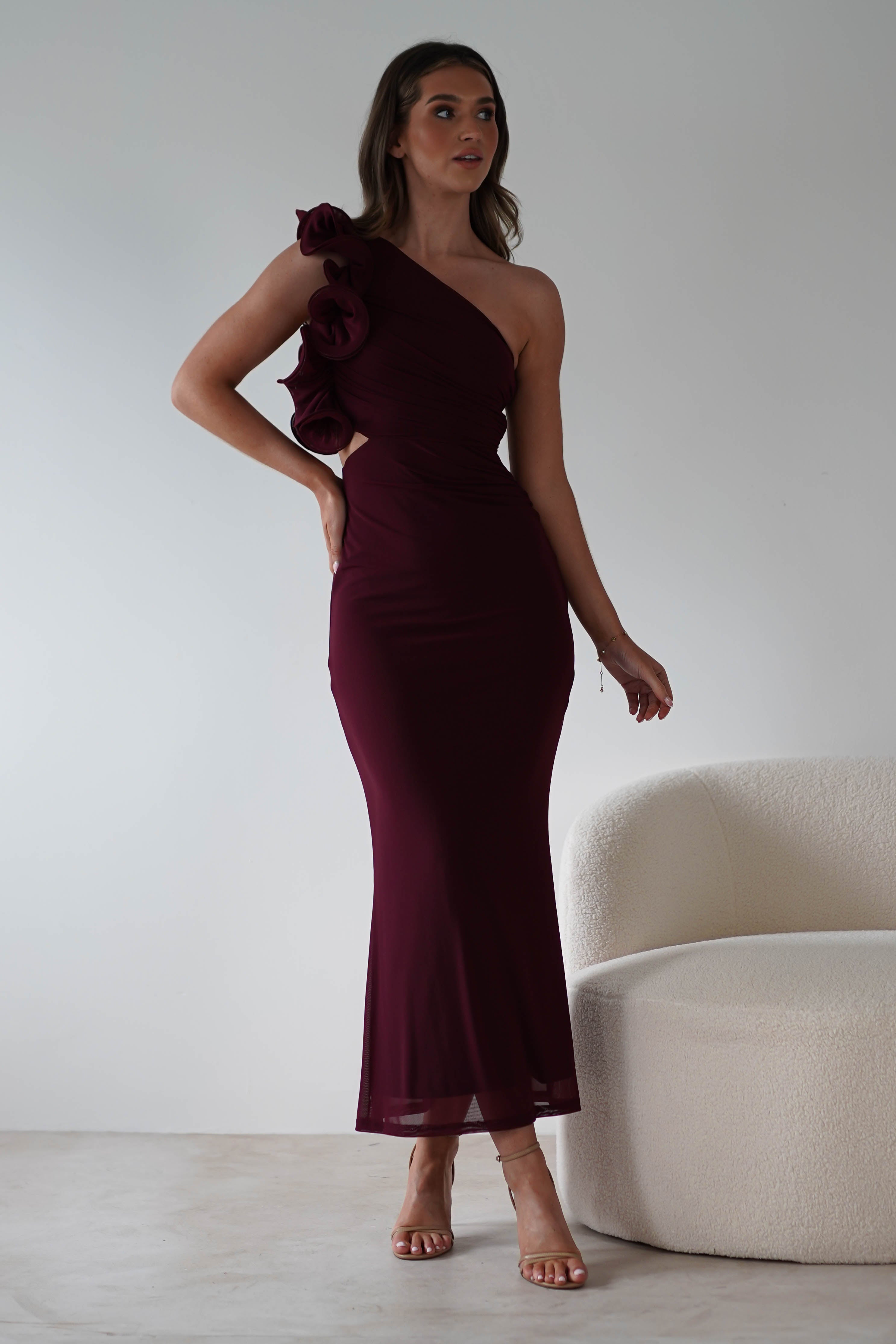 A woman in a fitted, one-shoulder dark burgundy midaxi dress with a ruffle feature stands confidently near a light-colored textured sofa. The floor is light wood, and the wall is plain gray, creating a modern and elegant setting that speaks to wear all day comfort.