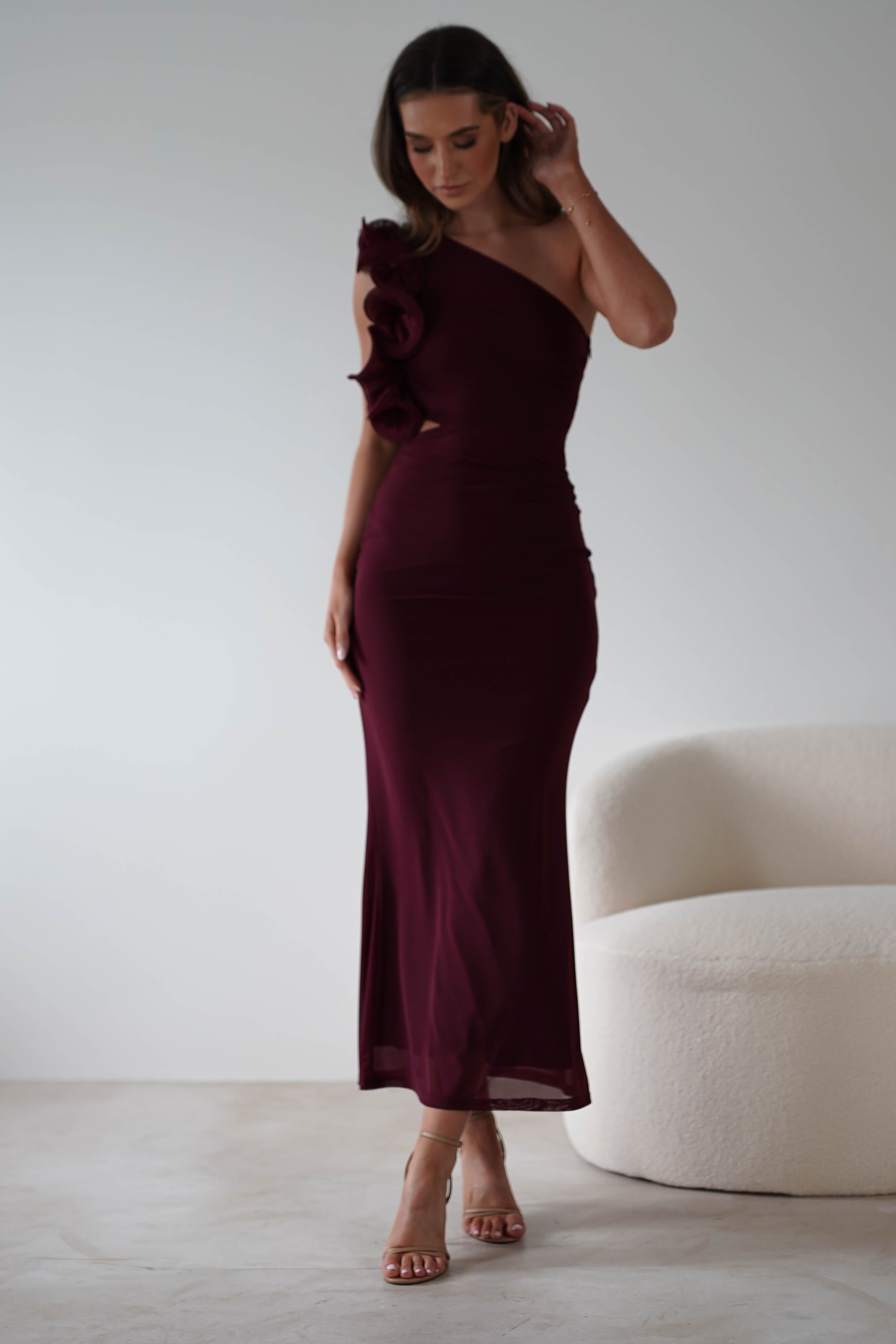 A woman in a burgundy one-shoulder midaxi dress with floral details and a subtle ruffle feature stands next to a white curved chair on a light-colored floor. Her long brown hair complements her strappy nude heels, offering wear-all-day comfort.