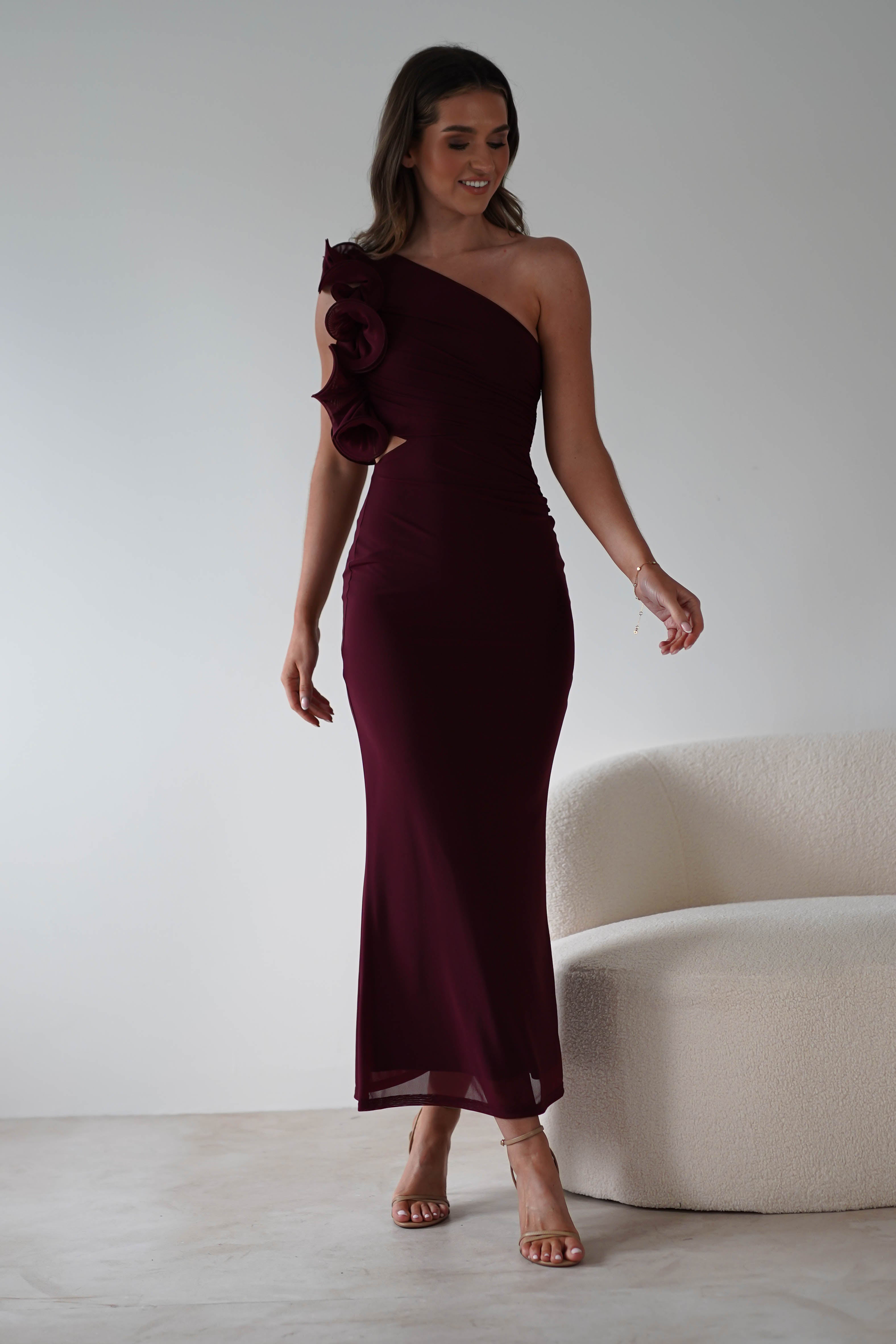 A woman in a burgundy midaxi dress with floral details on the shoulder stands next to a cream-colored sofa. She is wearing high heels and has long, wavy hair. The ruffle feature of her gown adds elegance against the plain white wall.