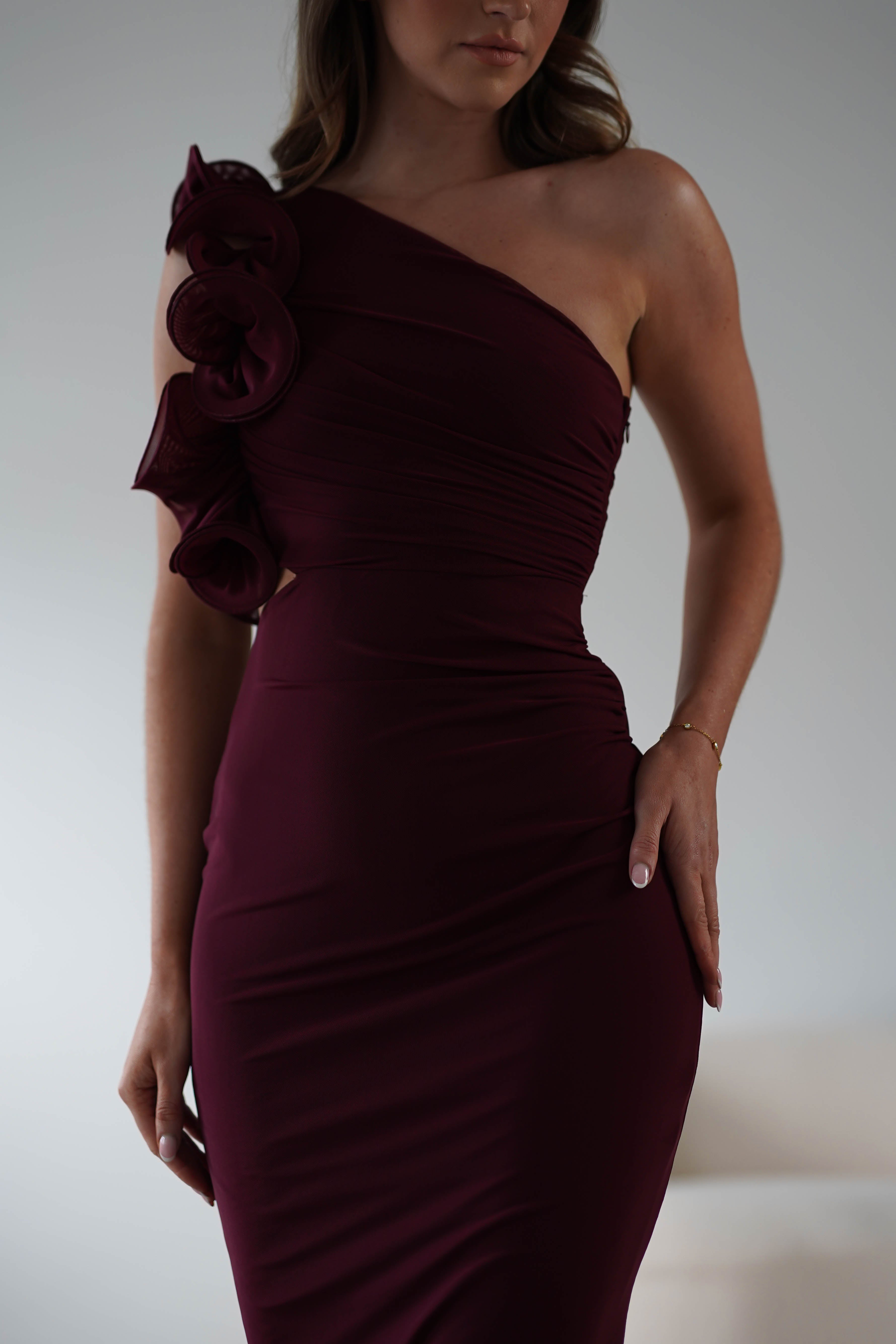 A person in a strapless dark burgundy midaxi dress with floral ruffle features on the shoulder poses against a white background. They have one hand on their hip and the other hanging down, exuding elegance and wear-all-day comfort.