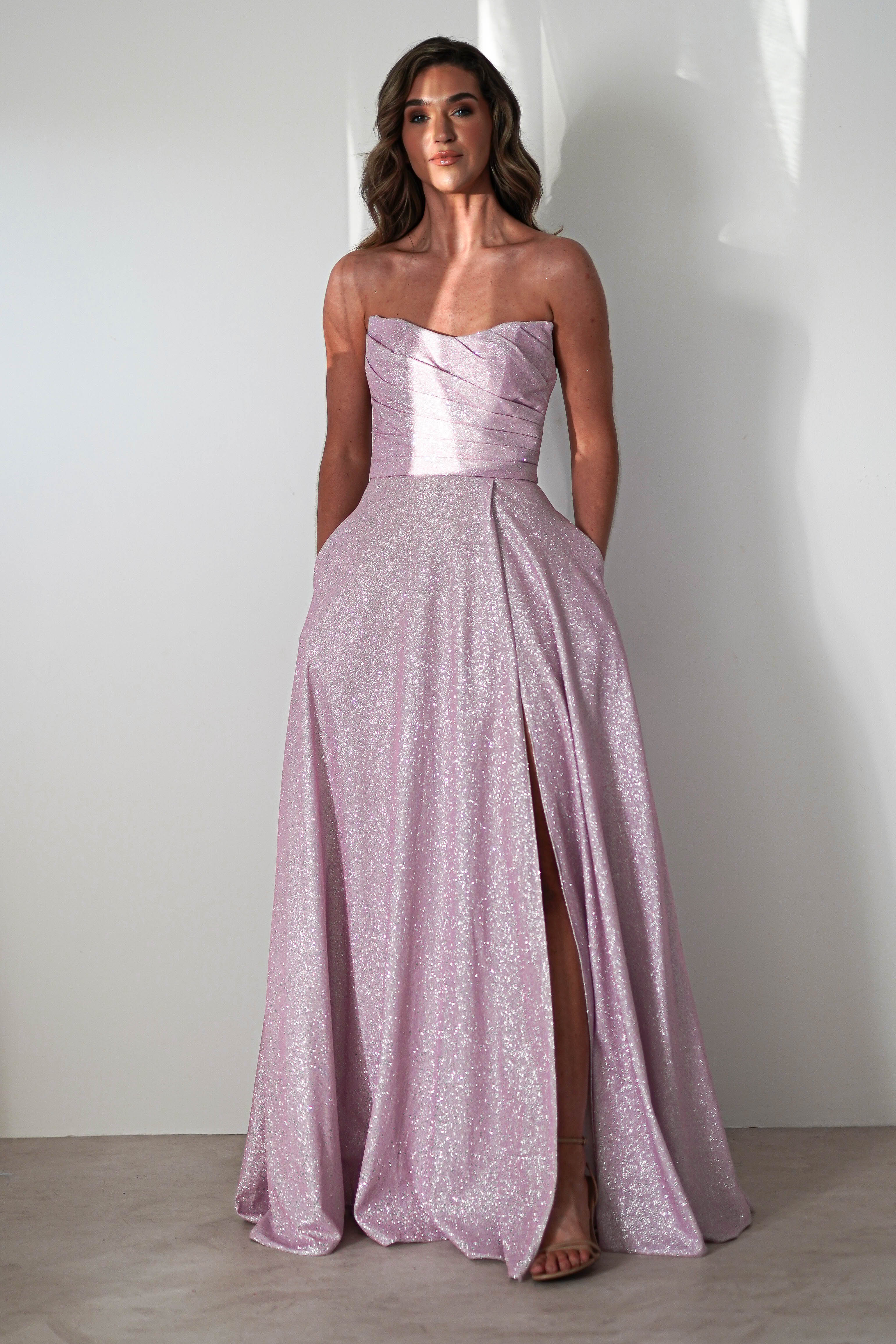 A woman in a strapless, light pink prom dress with a glittery finish and A-line silhouette stands against a plain white wall. The gown features a structured bodice and high slit. She has long, wavy hair and is wearing high heels, her hands tucked casually into the gowns pockets.