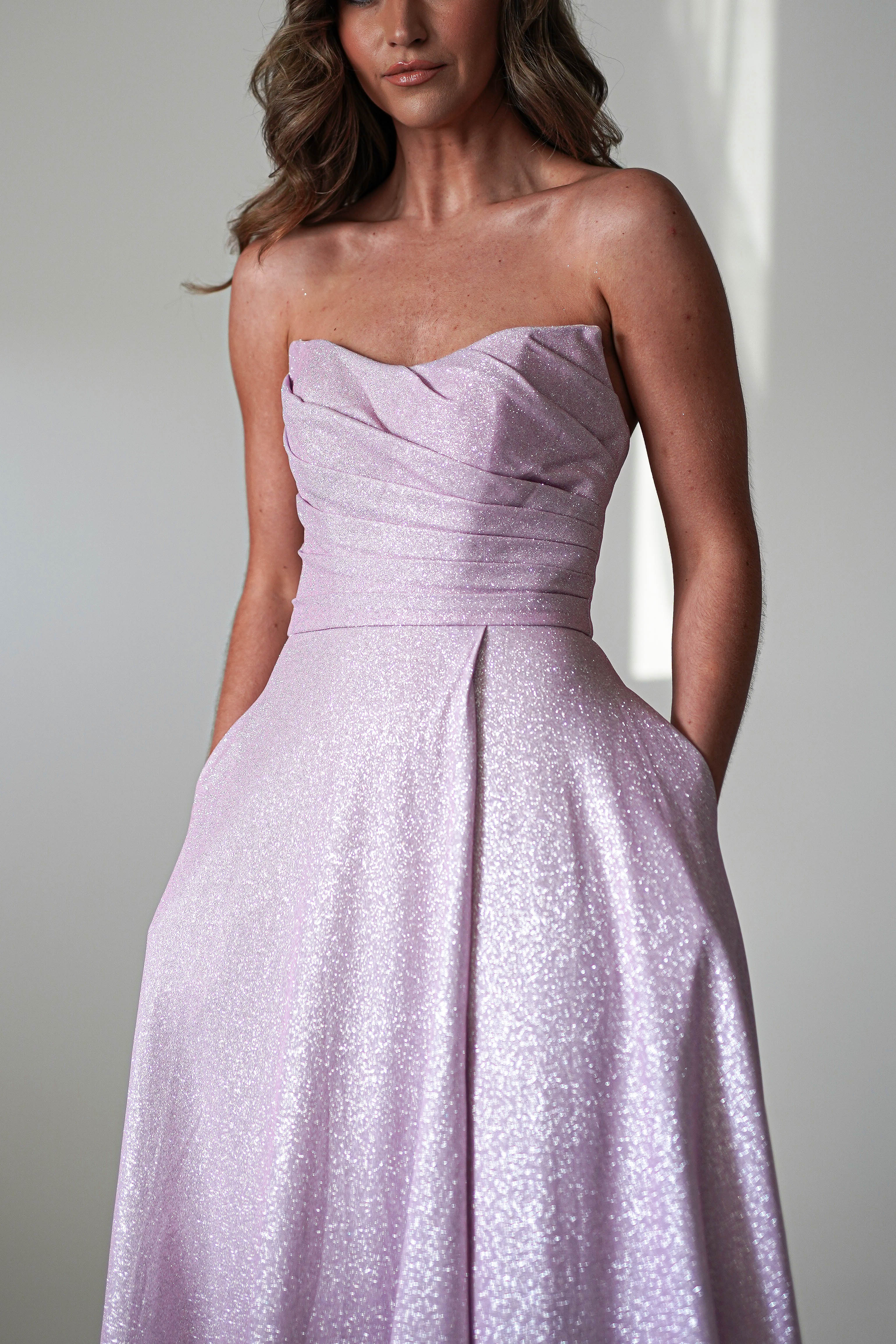 A person wearing a sleeveless, shimmering lavender evening gown with a fitted bodice and full skirt. The dress, perfect as a prom dress, features an A-line silhouette with textured fabric and pleated design. The background is softly lit in neutral tones.