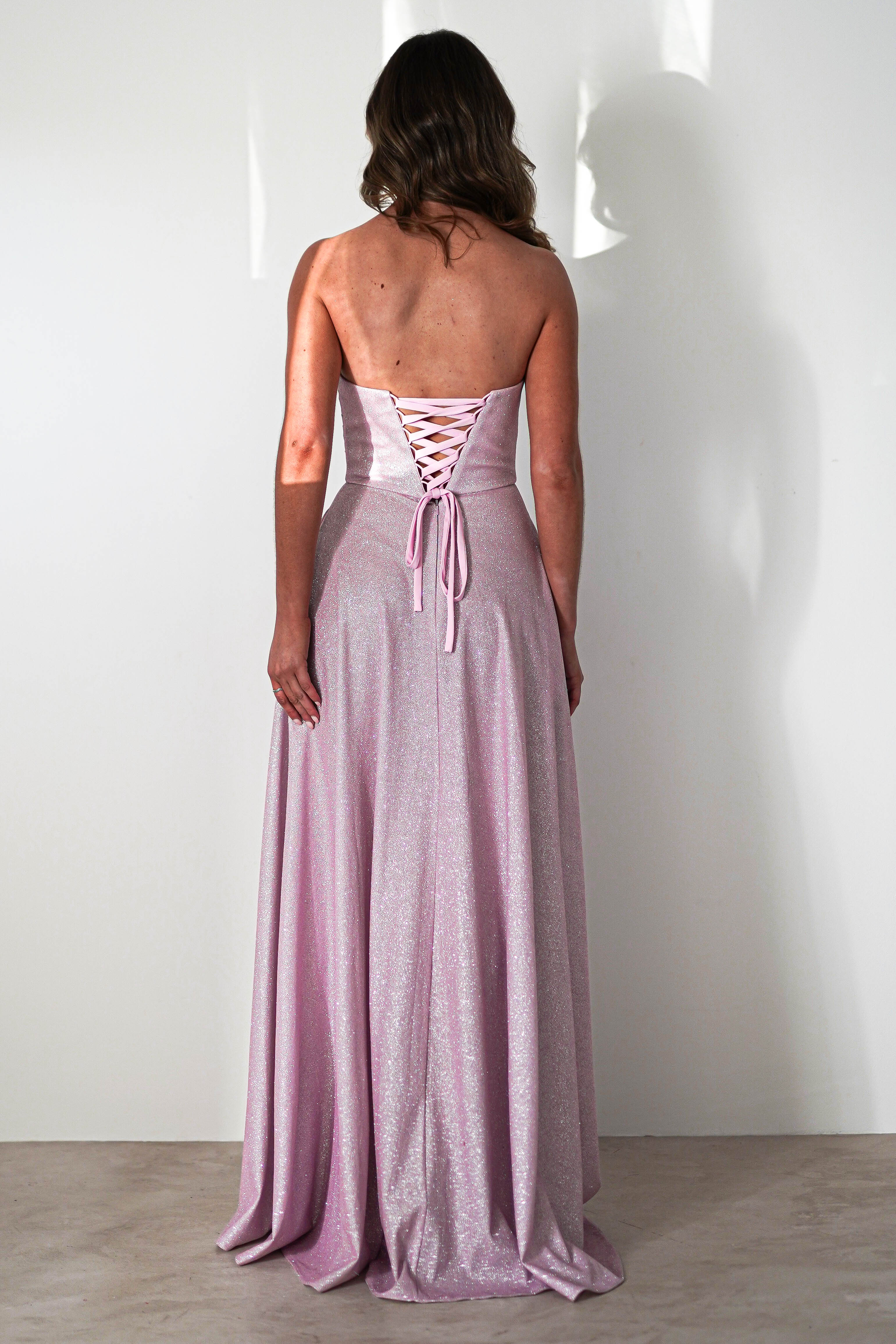 A woman with brown hair stands facing a white wall, wearing a sparkly pink strapless prom dress with a lace-up back. The A-line silhouette flows elegantly to the floor. The lighting casts a soft shadow.