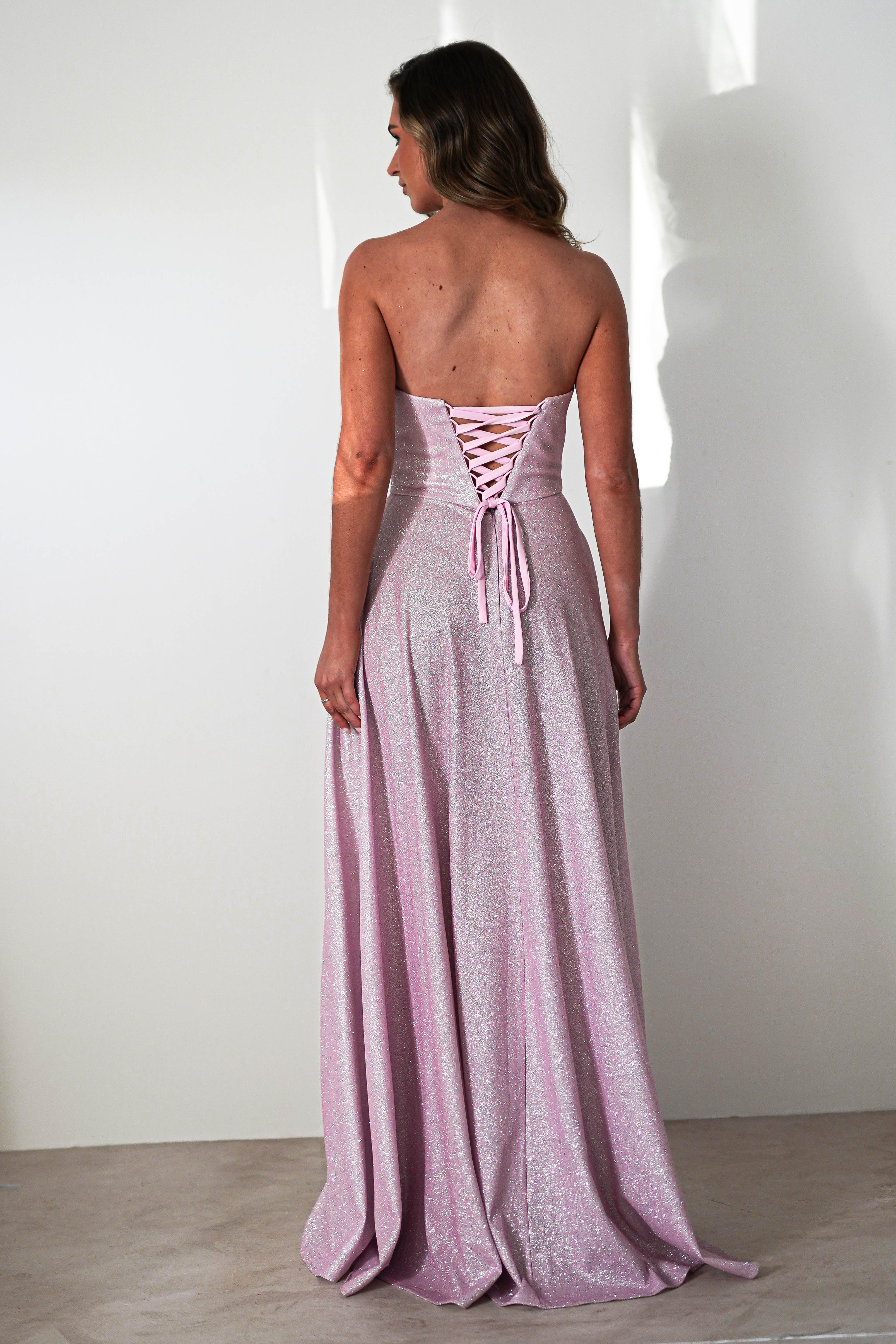 Rear view of a person wearing a sparkly pink strapless prom dress with a lace-up back. The A-line silhouette gracefully flows into a floor-length skirt. They stand against a plain white background, exuding elegance and charm.