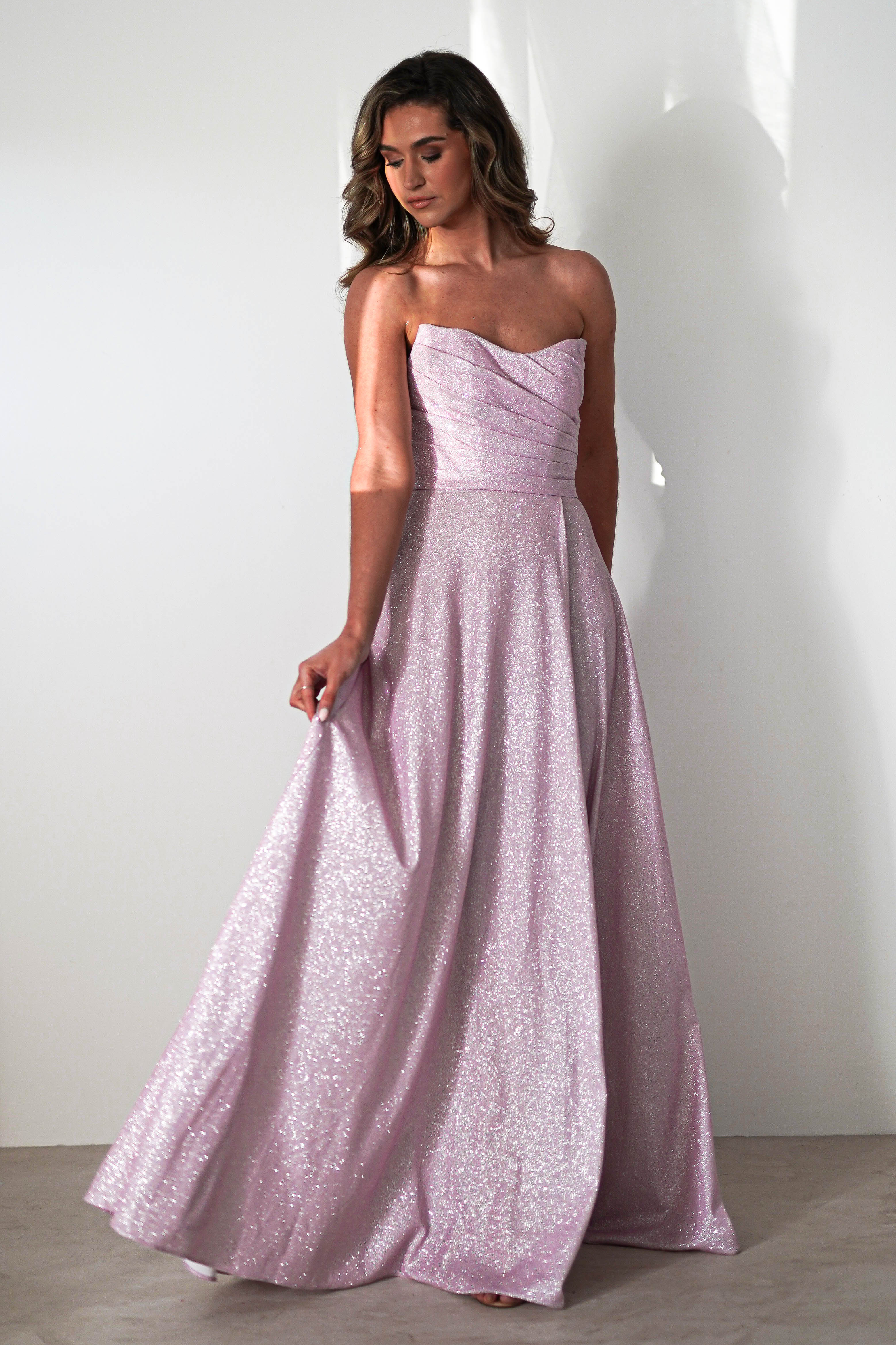A woman in a strapless, shimmery pink evening gown stands gracefully against a plain white background. She holds the gown slightly out to the side, enhancing its flowy A-line silhouette. Her long hair cascades elegantly over her shoulder.