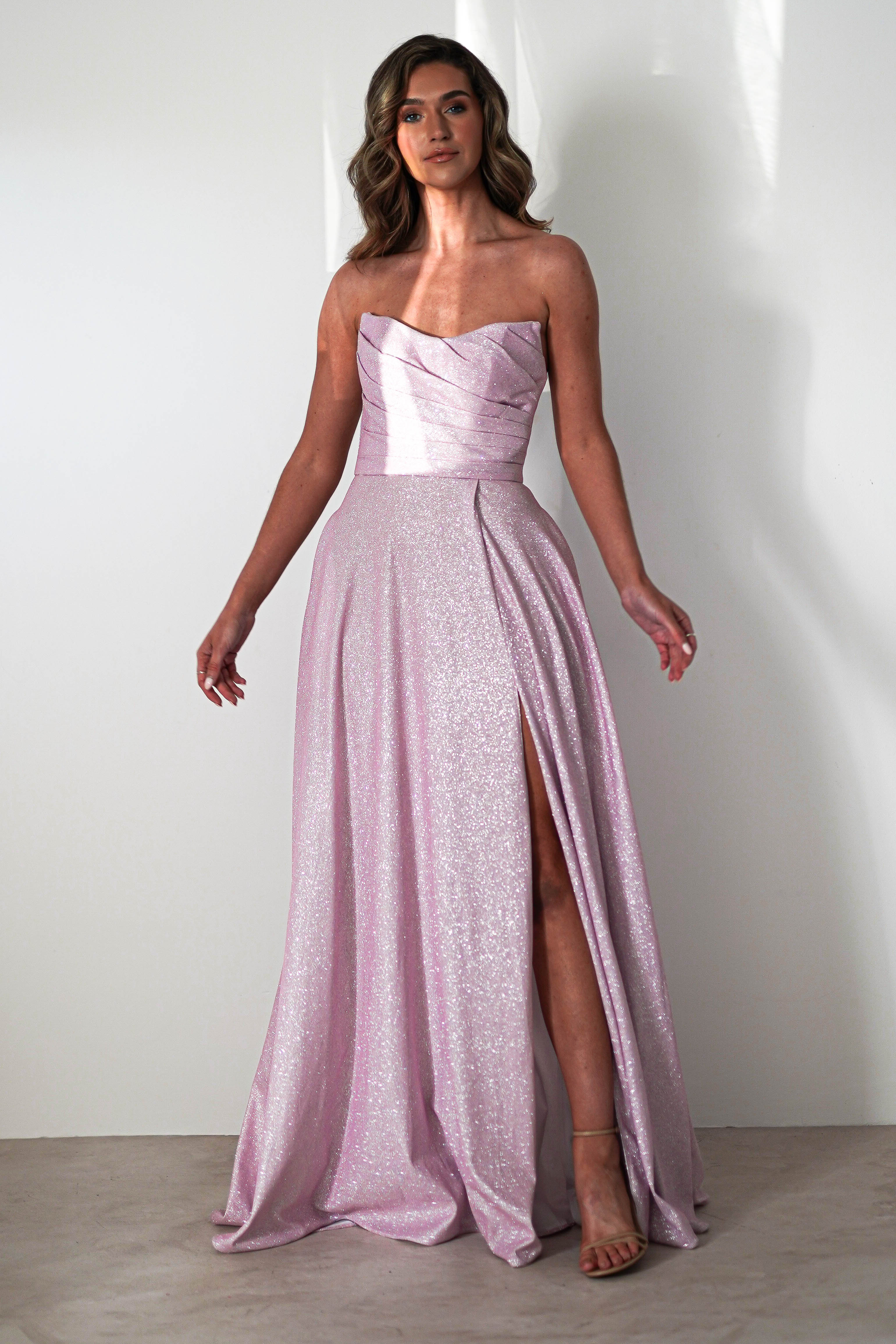 A woman in a strapless, shimmering pink prom dress with a sweetheart neckline and high slit stands against a white background. The A-line silhouette beautifully complements her wavy hair and high heels.