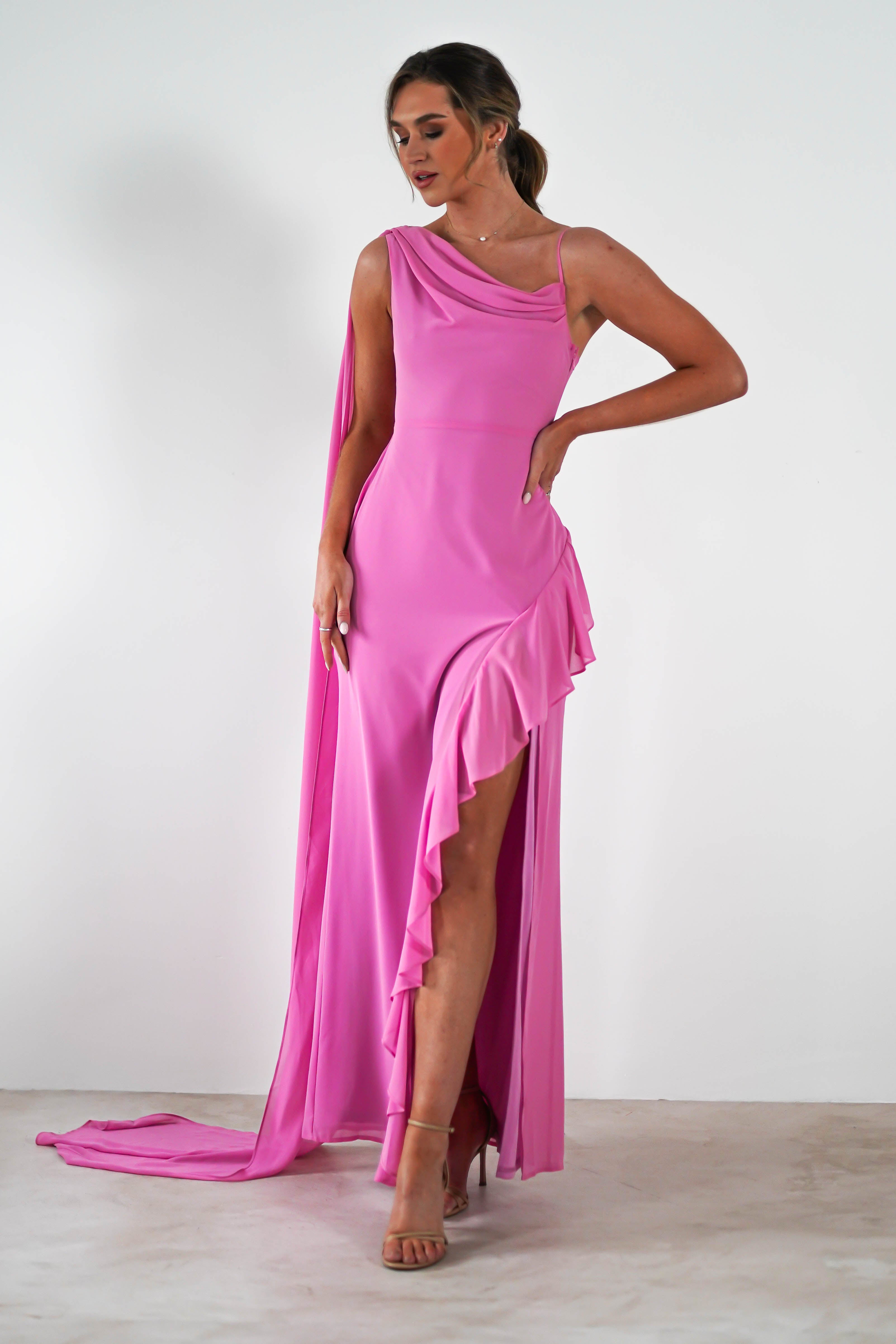 Woman in a sleeveless, coral one-shoulder gown with a thigh-high slit poses confidently. The floor-length dress features draped fabric and ruffled detail. She stands against a plain white background, wearing high heels, perfectly dressed as an elegant wedding guest.