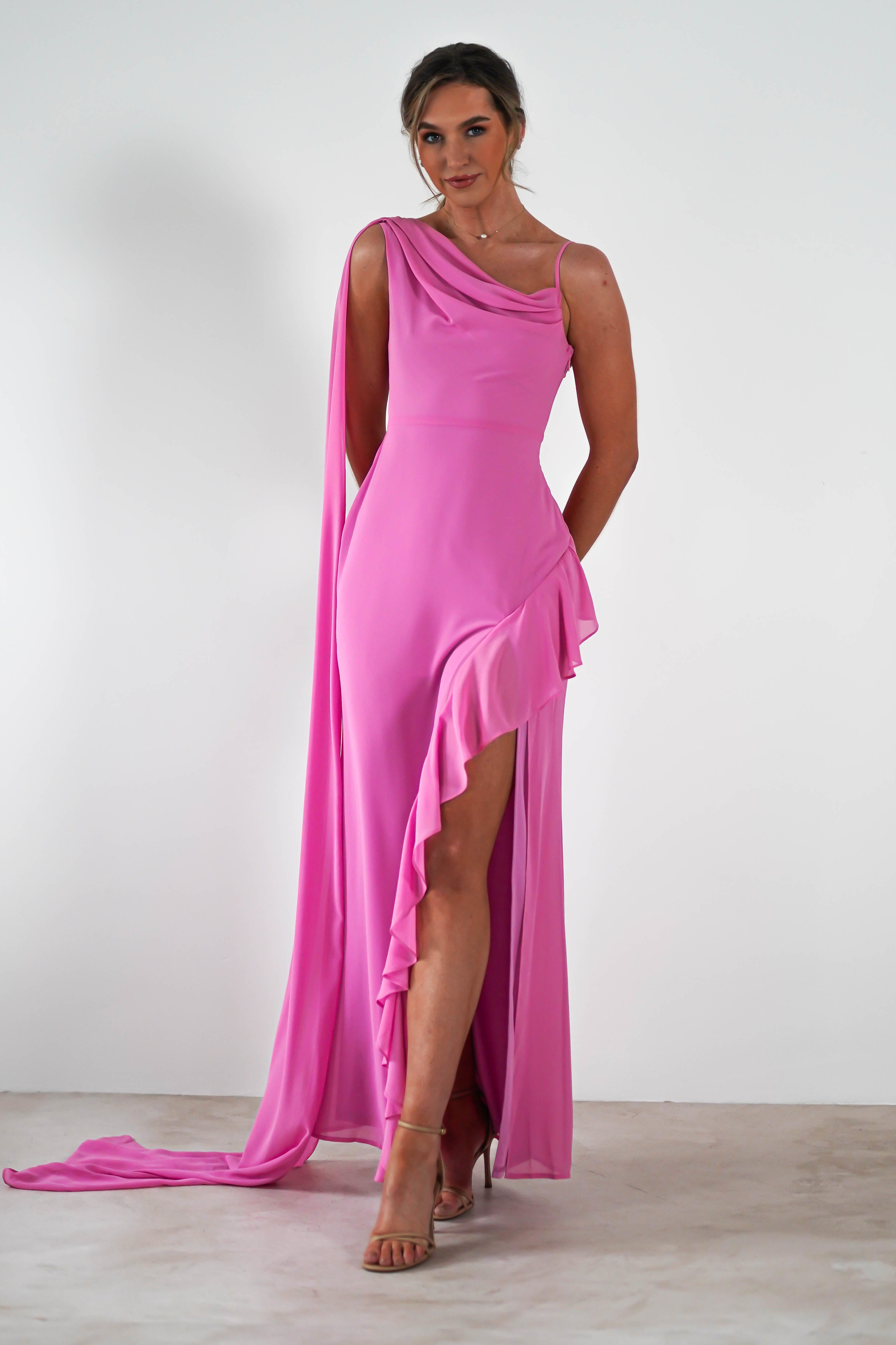 A person poses against a plain background, wearing a bright pink, sleeveless gown with an elegant draped neckline and a ruffled slit. They have light hair styled up and are wearing strappy high heels.