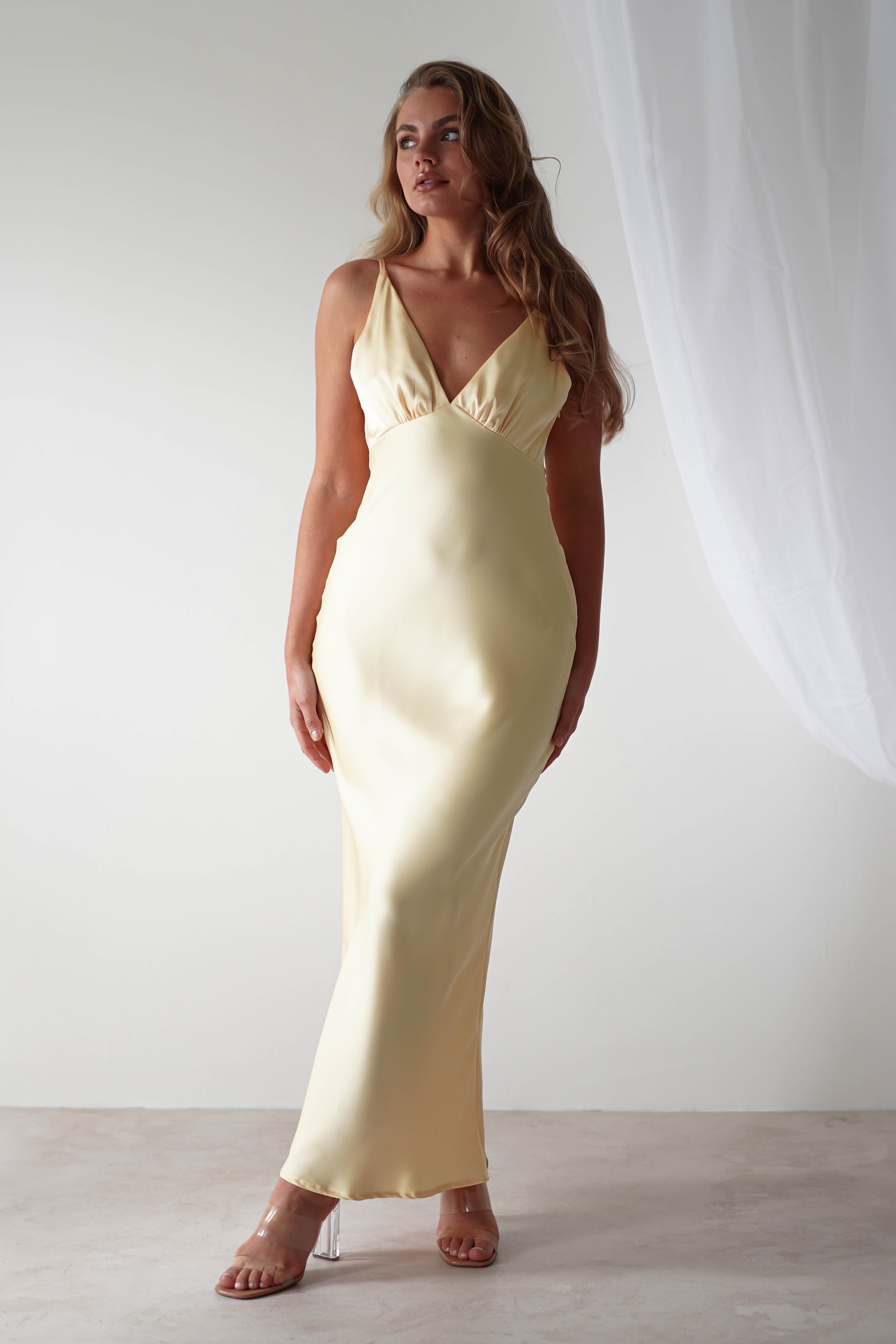 A person with long hair is wearing a sleek, light yellow satin maxi dress featuring a deep V-neckline. They are standing on a light-colored floor with a white curtain in the background. The setting is minimal and elegant, perfect for a graceful wedding guest appearance.