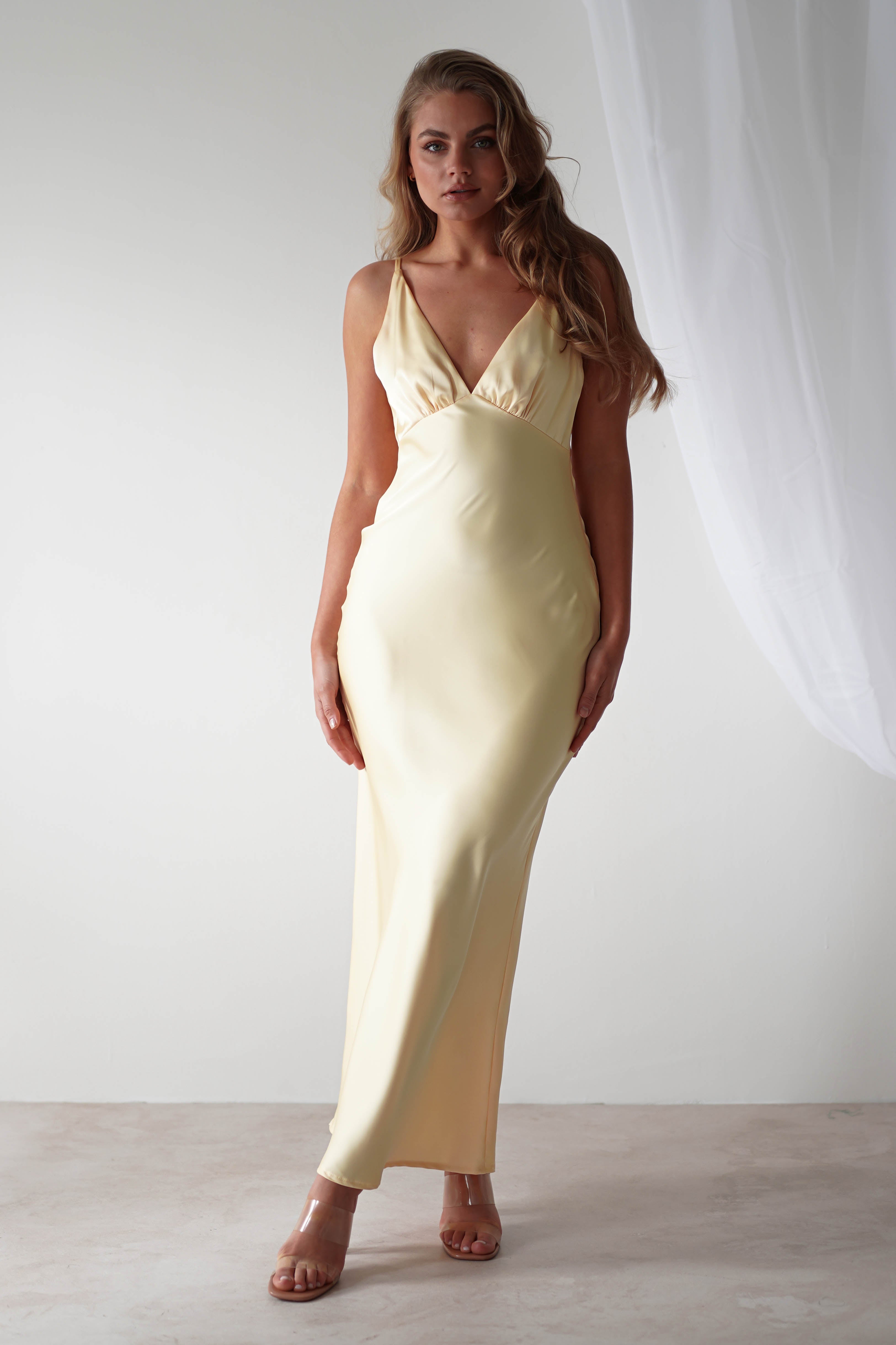 A woman in a long, elegant, light yellow satin maxi dress stands in a minimalistic setting with a white backdrop and soft lighting. She has long, wavy hair and is wearing strappy heels. One curtain is partially visible on the right.