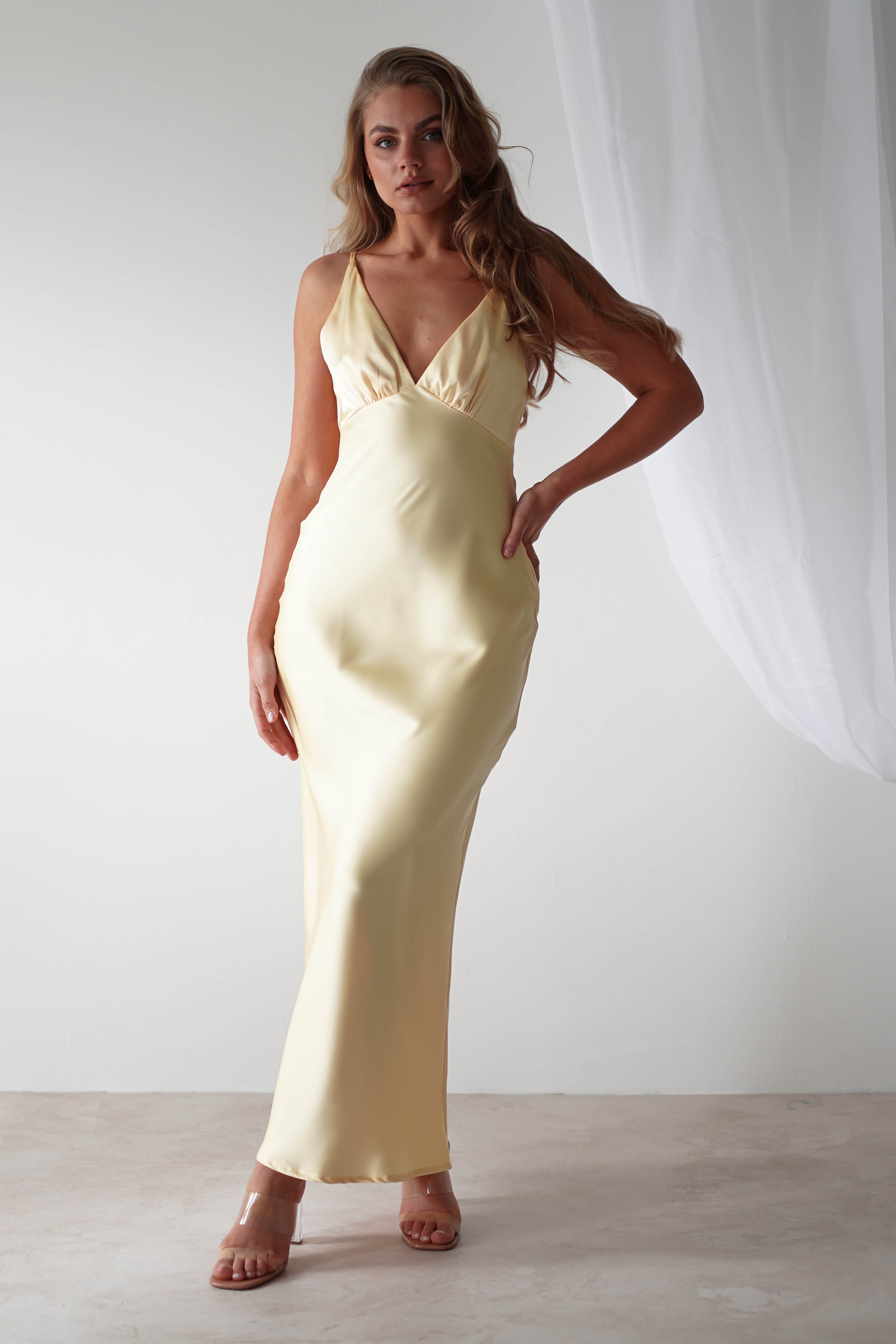 A person poses in a long, elegant satin maxi dress in light yellow with a deep V-neckline. They stand on a neutral-colored floor with a sheer white curtain in the background, holding one hand on their hip—perfect as a wedding guest dress.