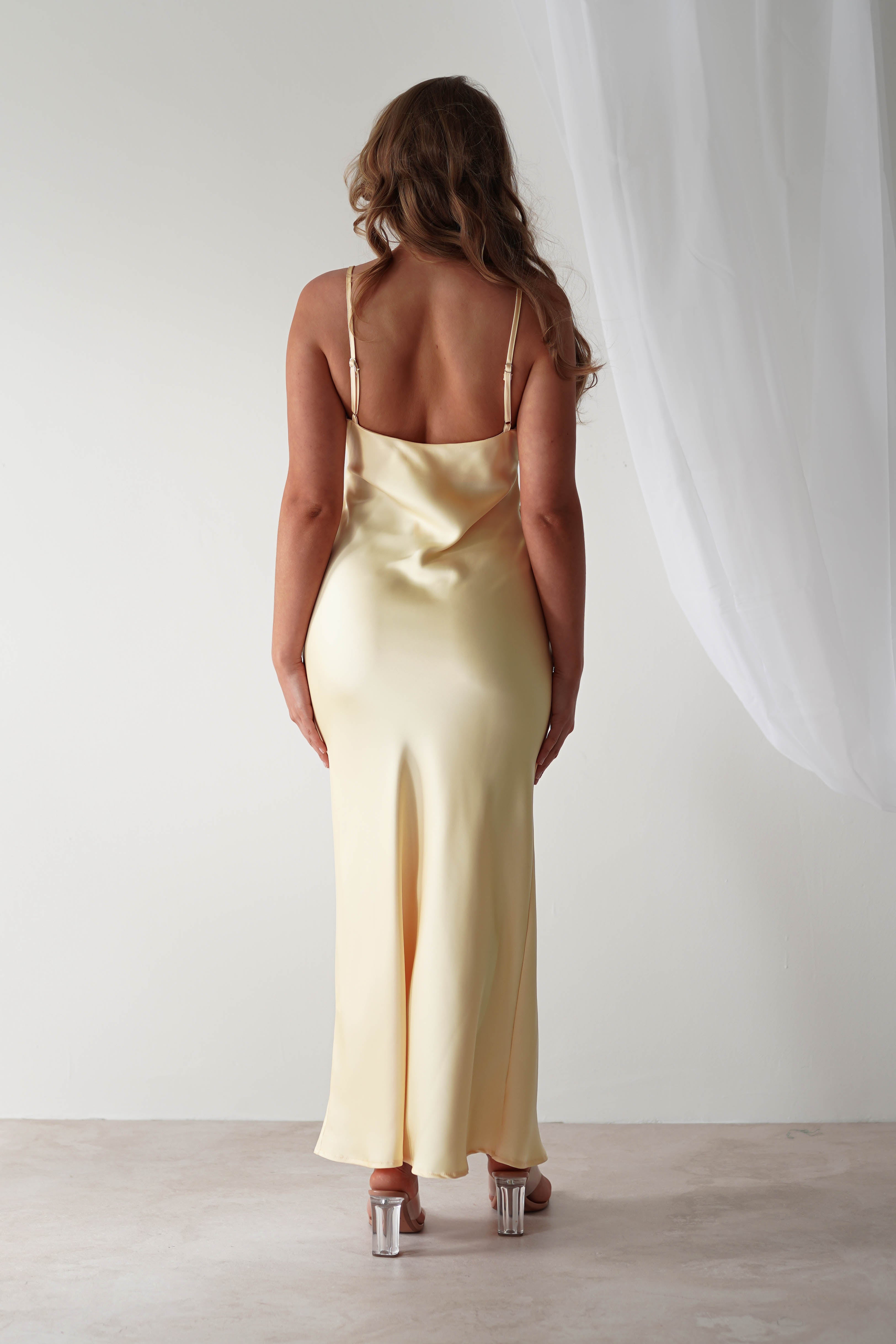 A woman stands with her back to the camera, wearing a light yellow satin maxi dress. The dress, perfect for a wedding guest, has thin straps and falls gracefully to the ankles. She stands on a neutral floor with a sheer white curtain hanging to the right.