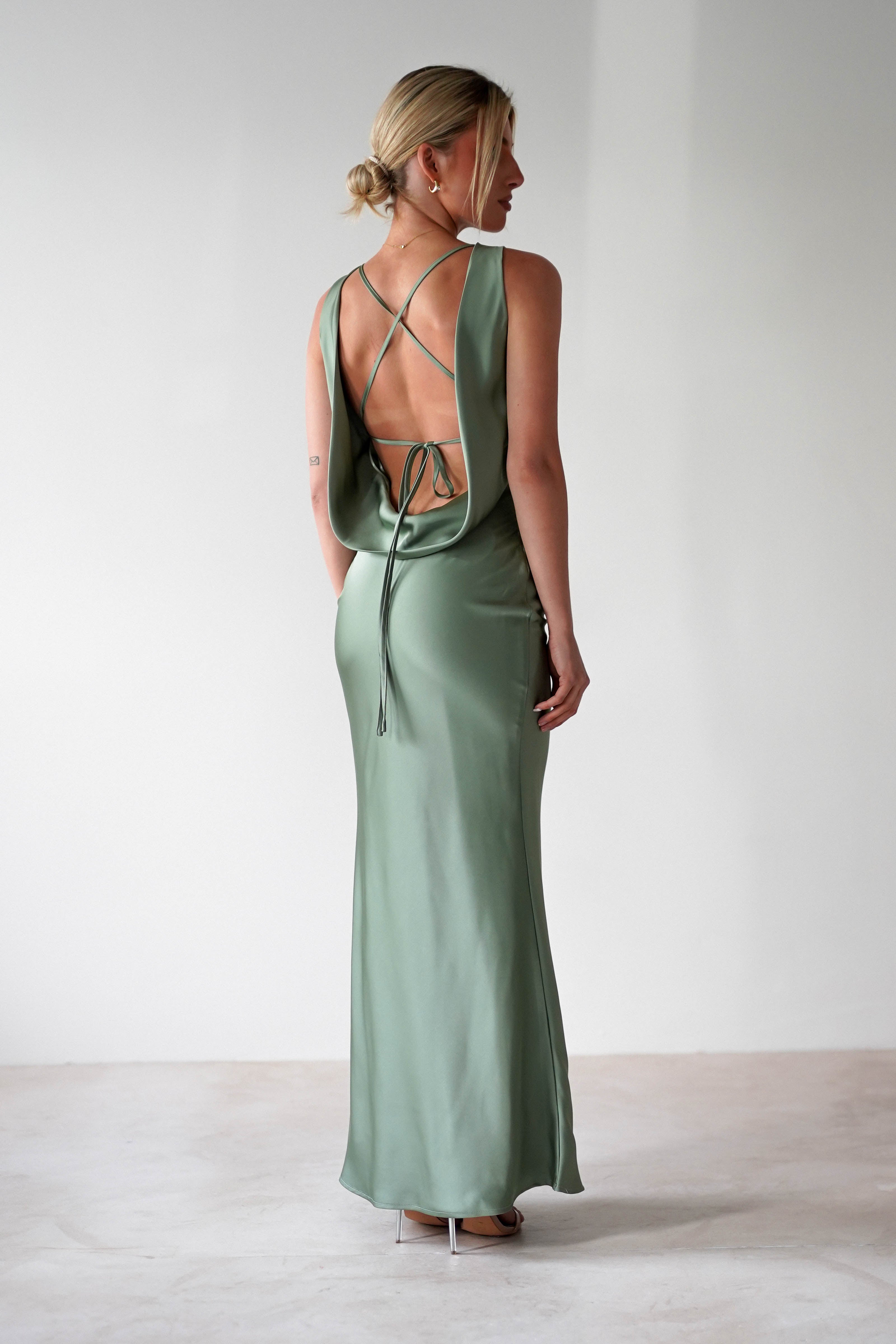 A woman stands on a light floor wearing a satin maxi dress in sage green with an open back and crisscrossed straps—perfect as a wedding guest dress. Her hair is in a bun, showcasing the dress’s elegant back detail.
